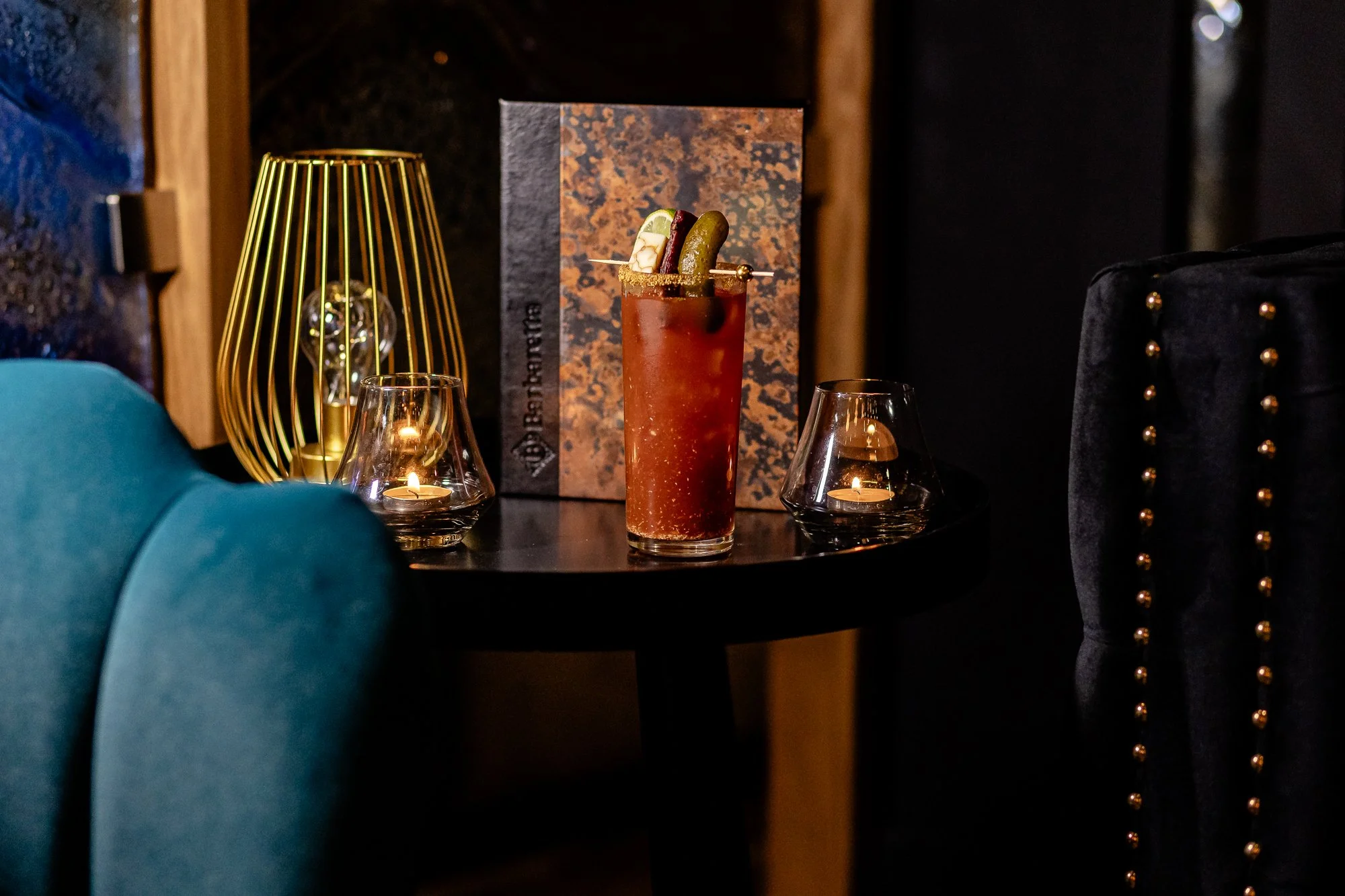 A tall cocktail glass with a red drink, garnished with lime, cherry, olive, and a pick, placed on a black table next to a candle holder with a lit candle, a decorative lamp, and a menu or sign in the background.