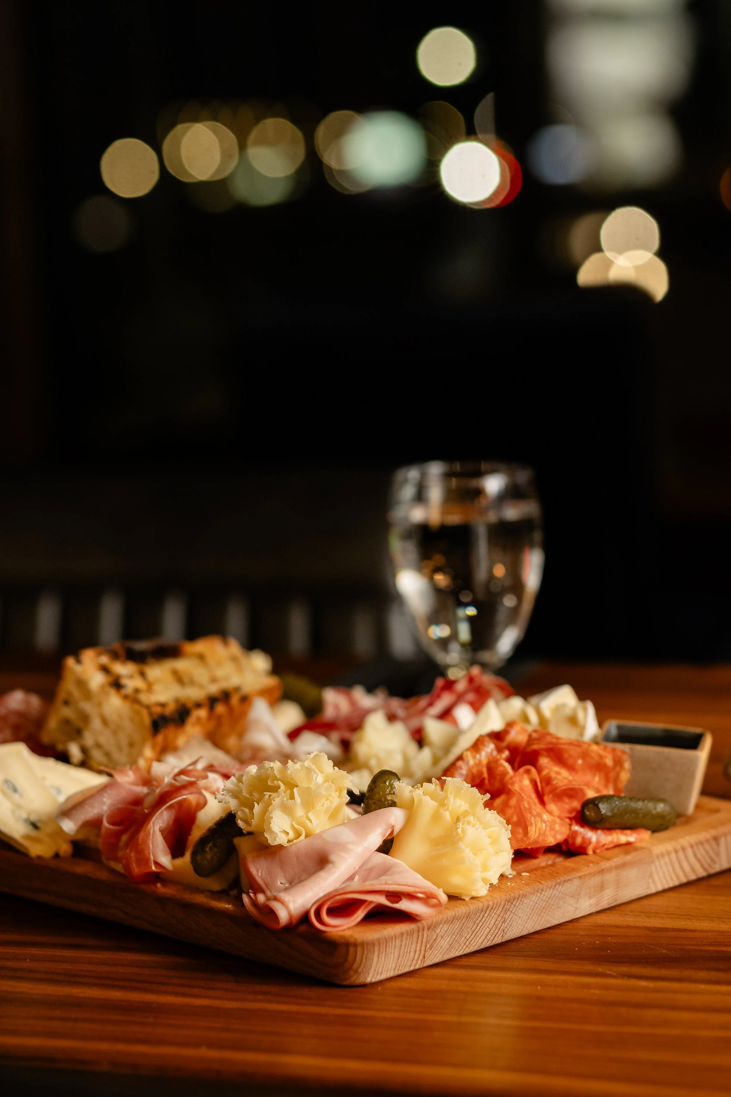 A cheese and charcuterie platter with assorted cheeses, meats, and pickles on a wooden board, with a glass of white wine in the background, out of focus.