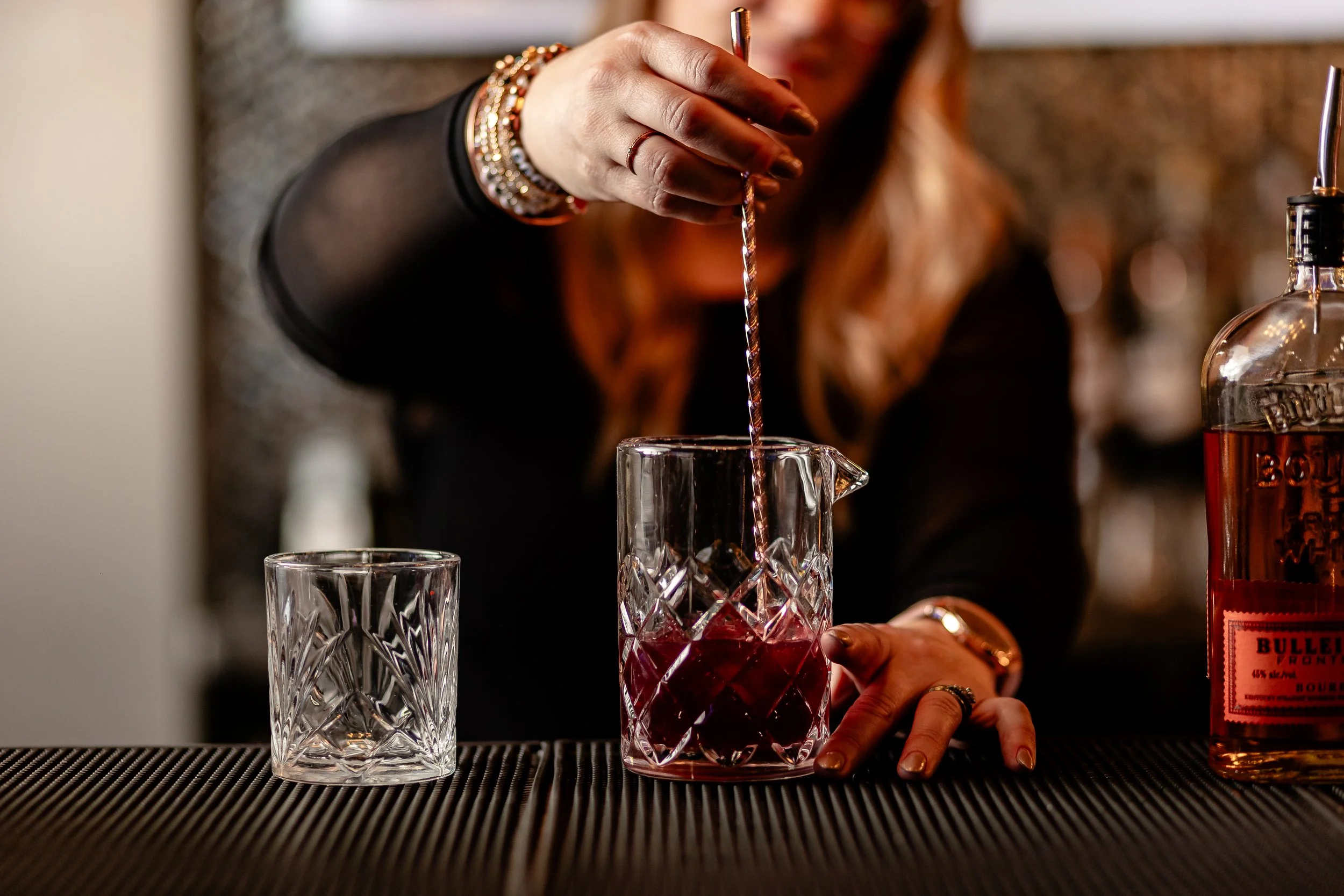 A person with multiple bracelets stirring a drink in a crystal glass with a twisted stirring stick, with a bottle of bourbon whiskey nearby and another empty crystal glass in the foreground.