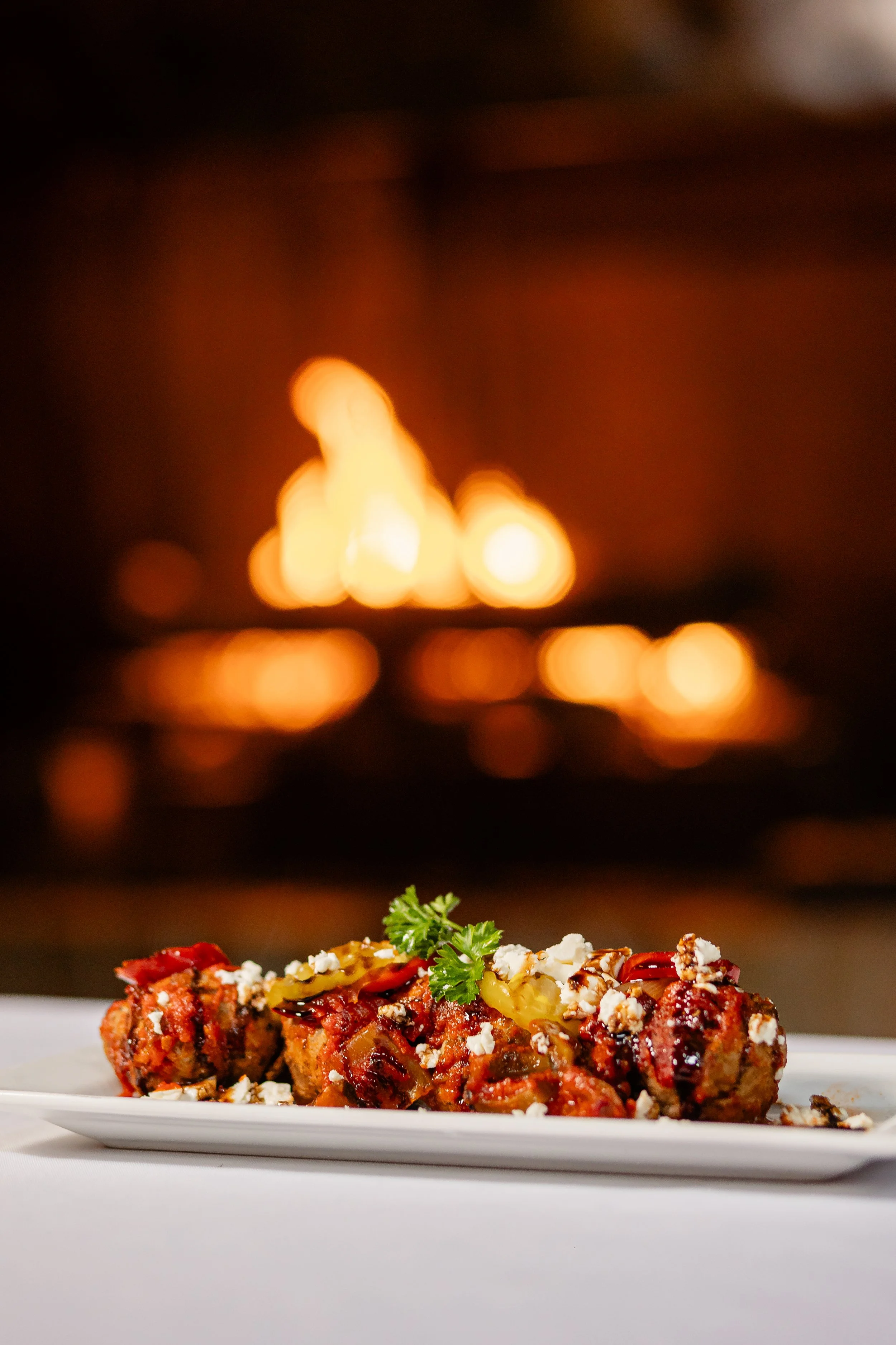 Close-up of a baked dish, possibly meatloaf, garnished with herbs and crumbled cheese, with a blurred fireplace in the background.