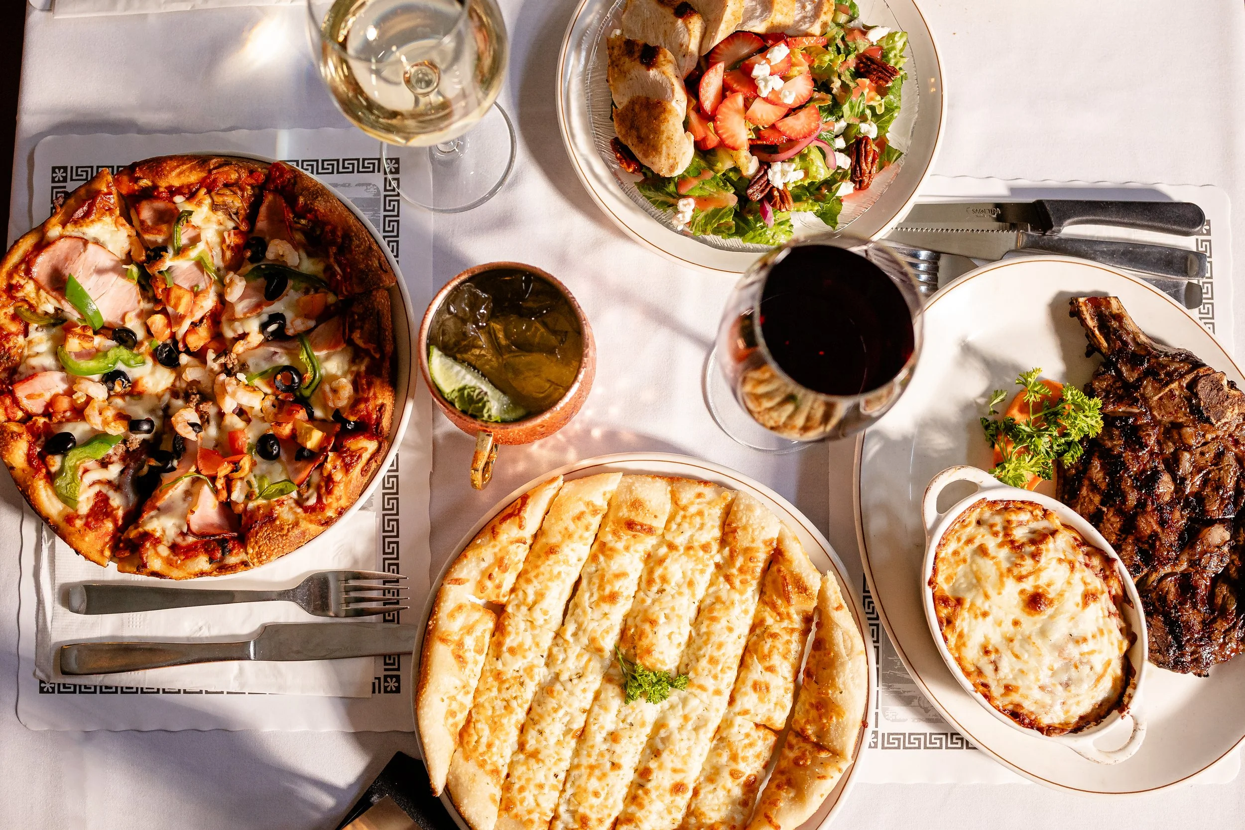 A variety of dishes on a white tablecloth, including pizza, salad, grilled meat, cheese bread, and drinks.