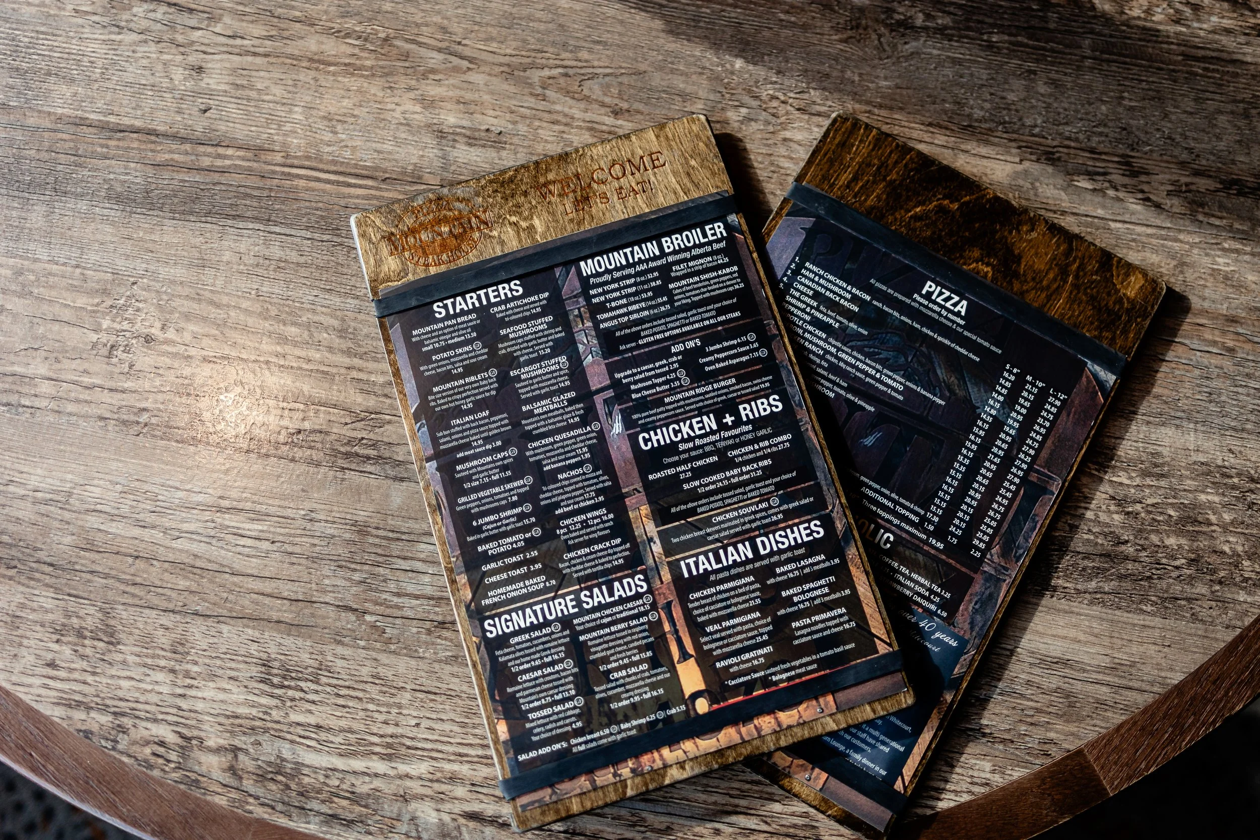 A wooden table with a black laminated menu featuring food and drink options, including starters, salads, pizzas, and ribs, from a restaurant.
