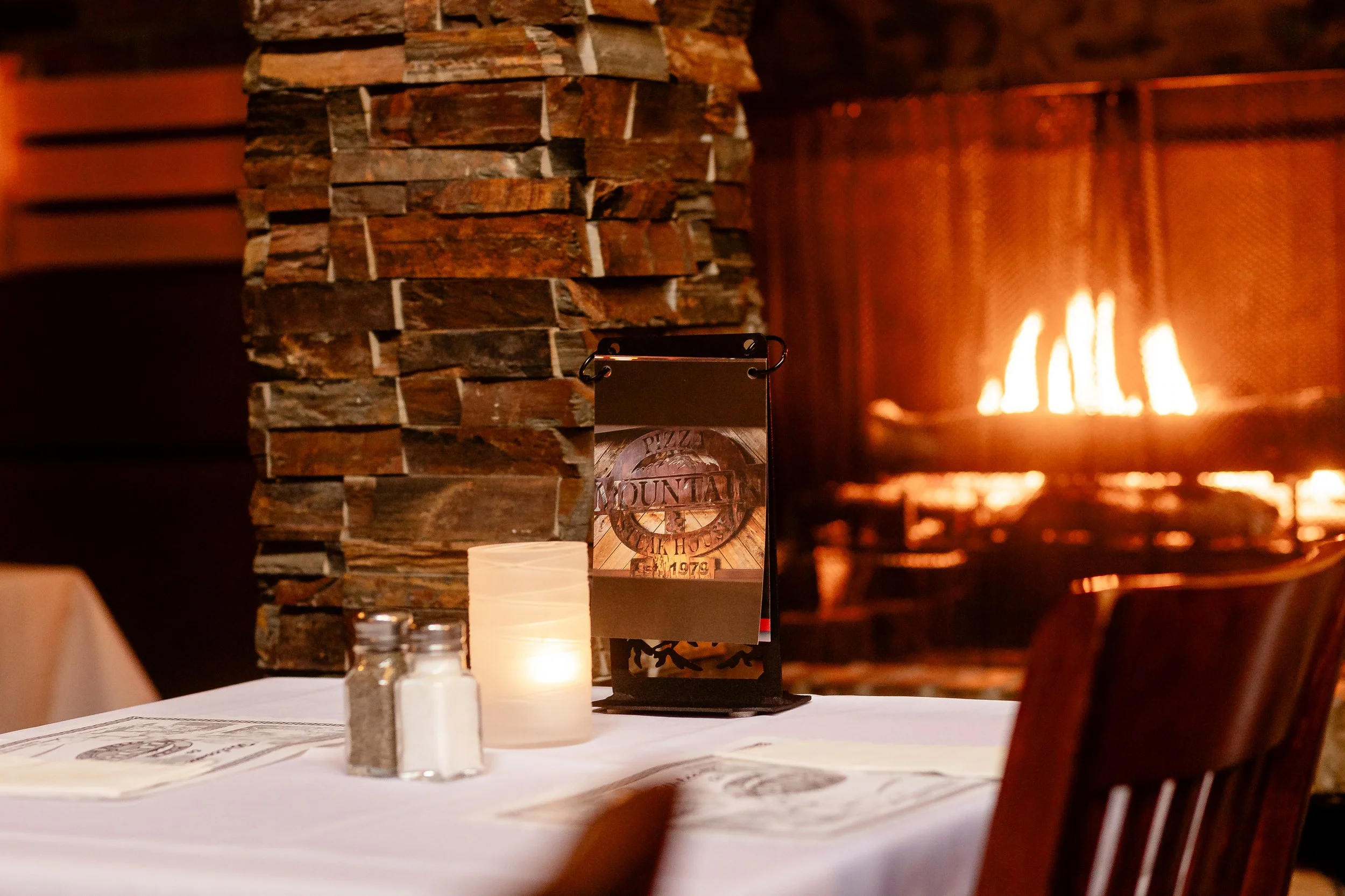 A cozy restaurant interior with a stone fireplace on the left, warm lighting, and a table set with candles, menus, and utensils. A sign or menu holder is on the table, and there's a visible wall with wooden paneling in the background.