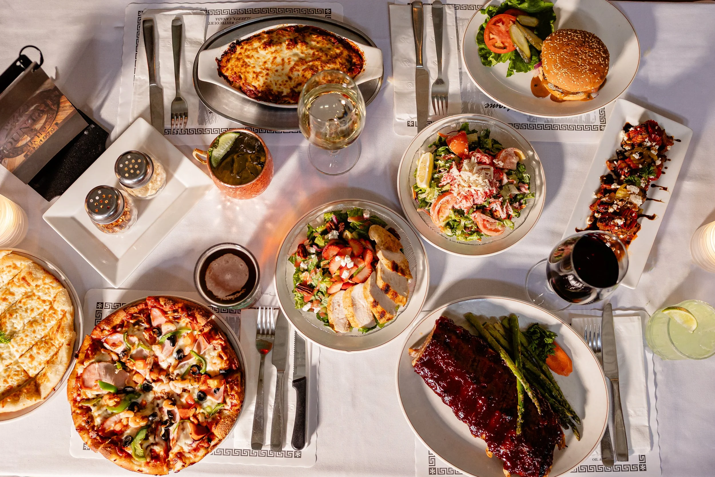 A variety of food dishes on a dining table, including pizza, salad, lasagna, grilled ribs with asparagus, a cheeseburger, and drinks, set on white tablecloths.