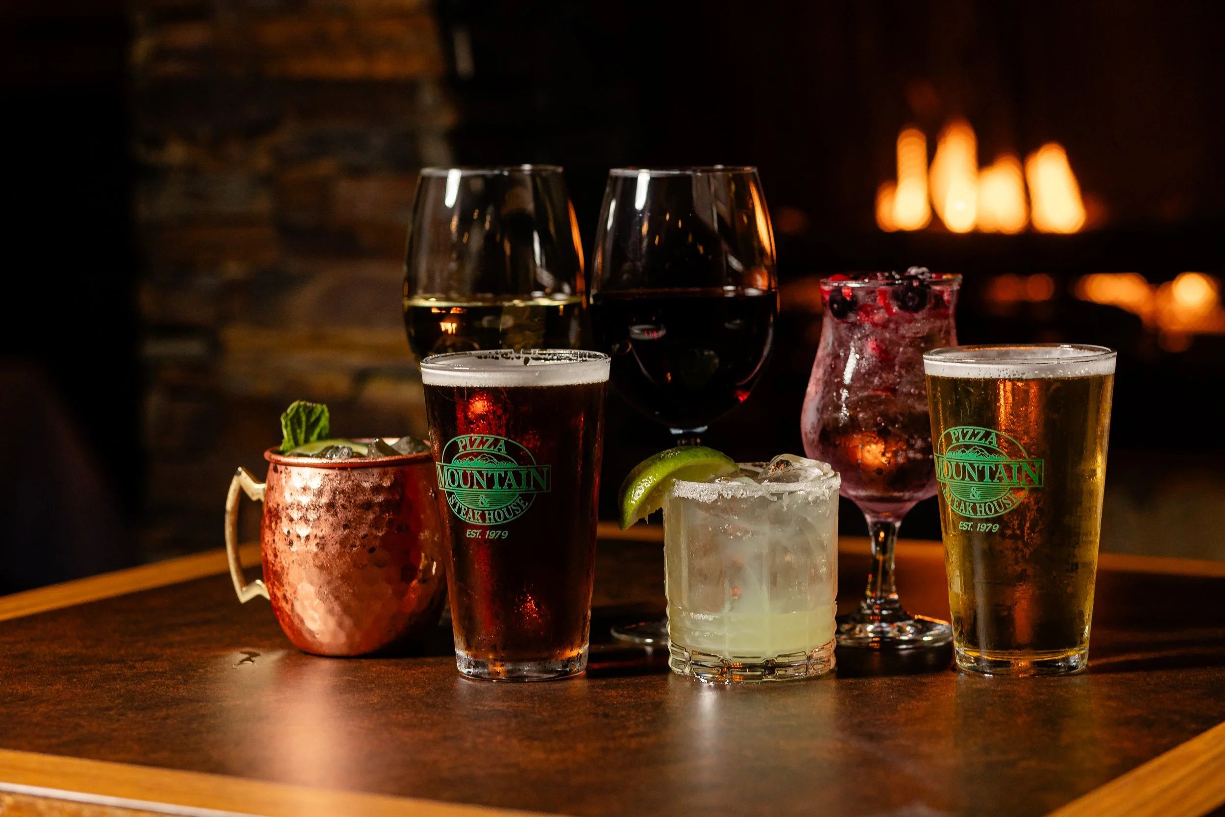 A variety of cocktails and drinks on a wooden table in front of a fireplace, including wine, beer, and mixed drinks with lime slices.