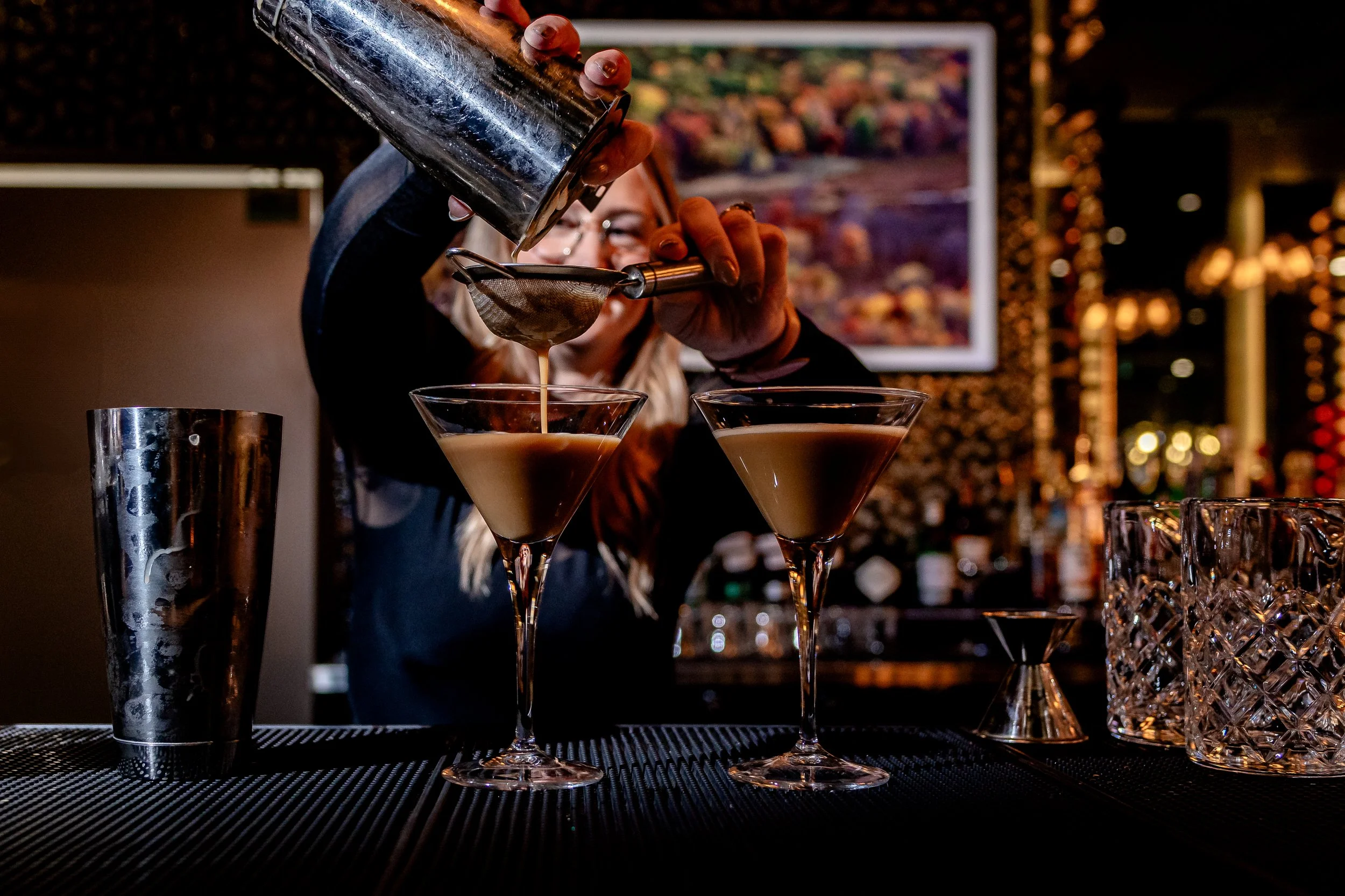 A bartender pours a cocktail through a strainer into martini glasses at a bar with a colorful background.