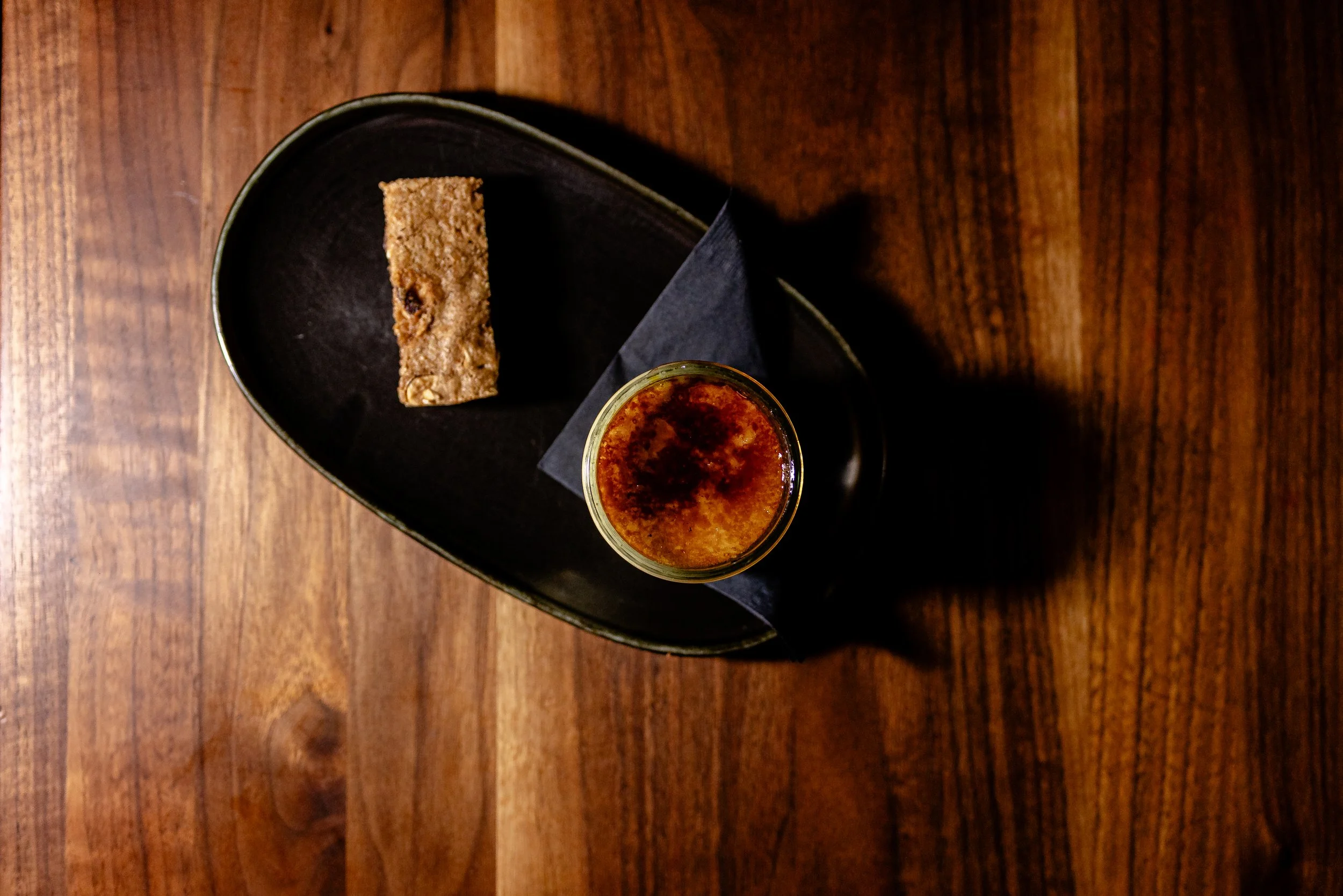 A glass of beer with a frothy head next to a granola bar on a black plate, on a wooden table.