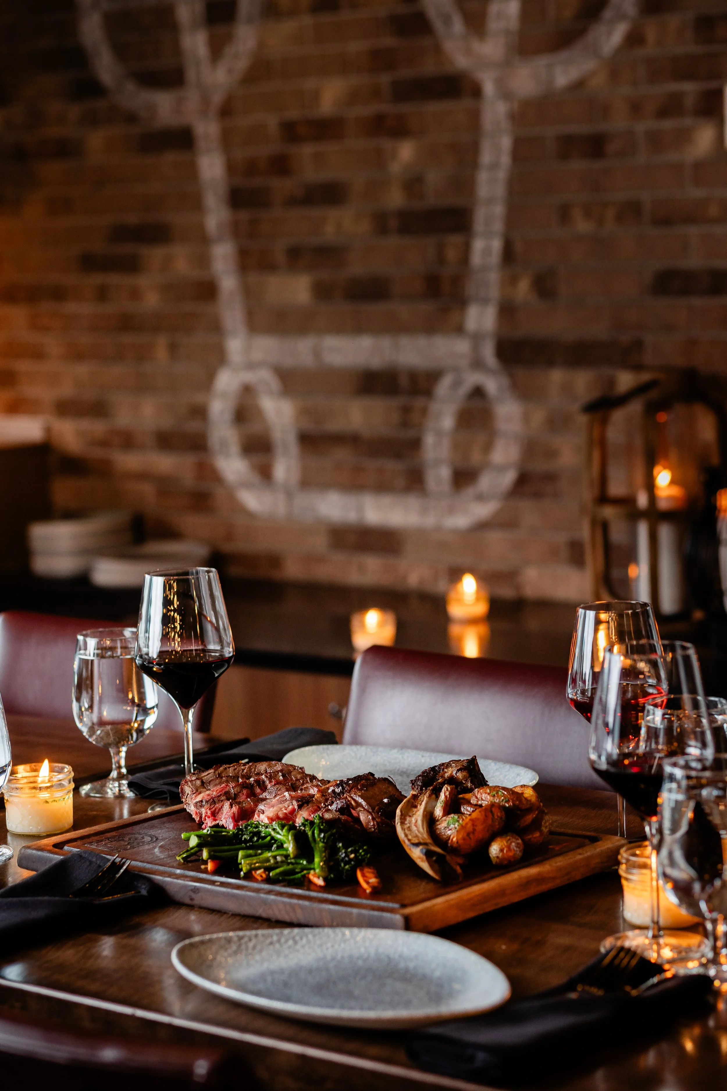 A dining table set for a meal with glasses of red wine, a candle, and a platter of steak, grilled vegetables, and green beans in a cozy, dimly lit restaurant with candles on the table and a brick wall with a large decorative horseshoe in the background.