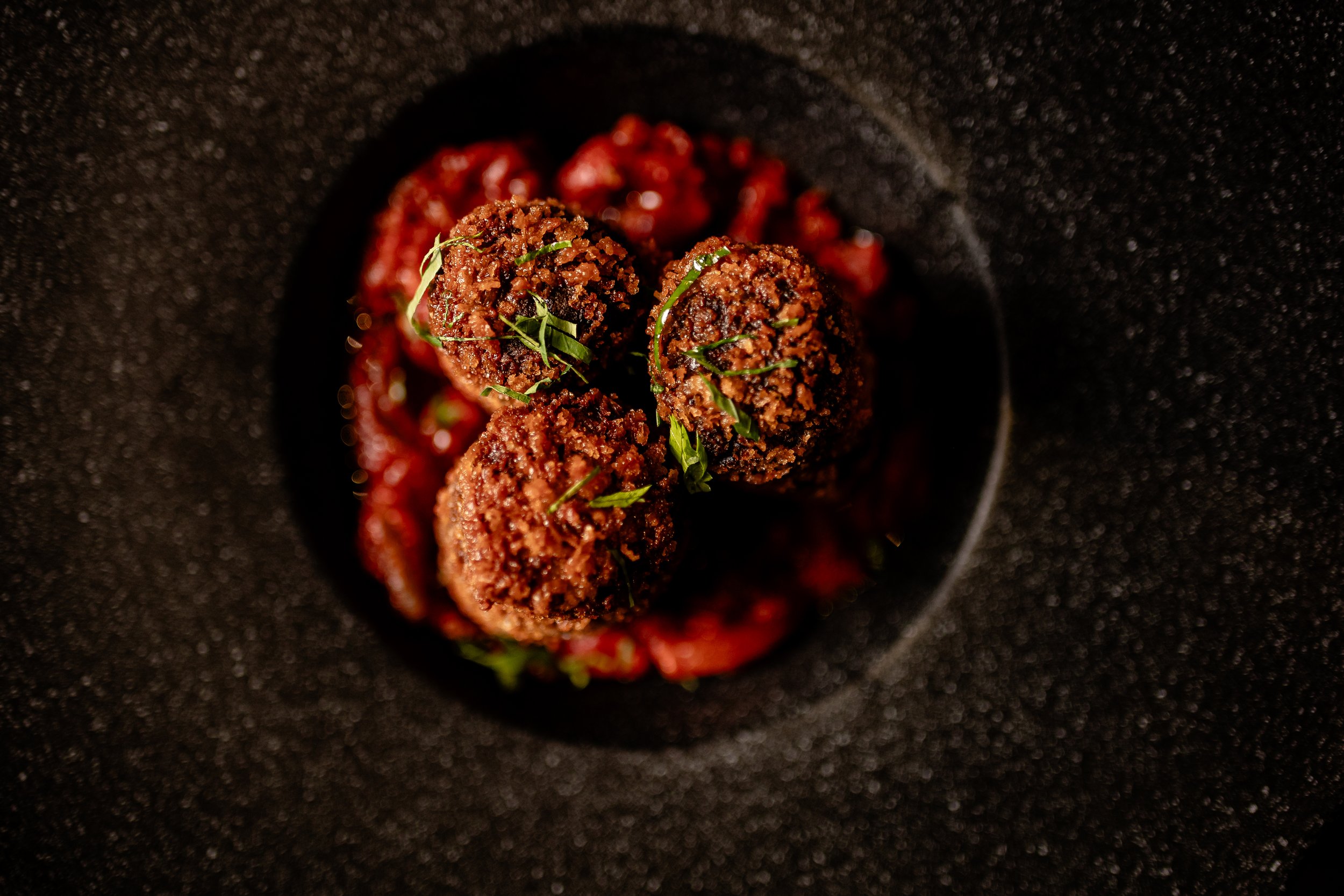 Close-up of three cooked meatballs garnished with chopped herbs on a black textured plate with a red sauce underneath.