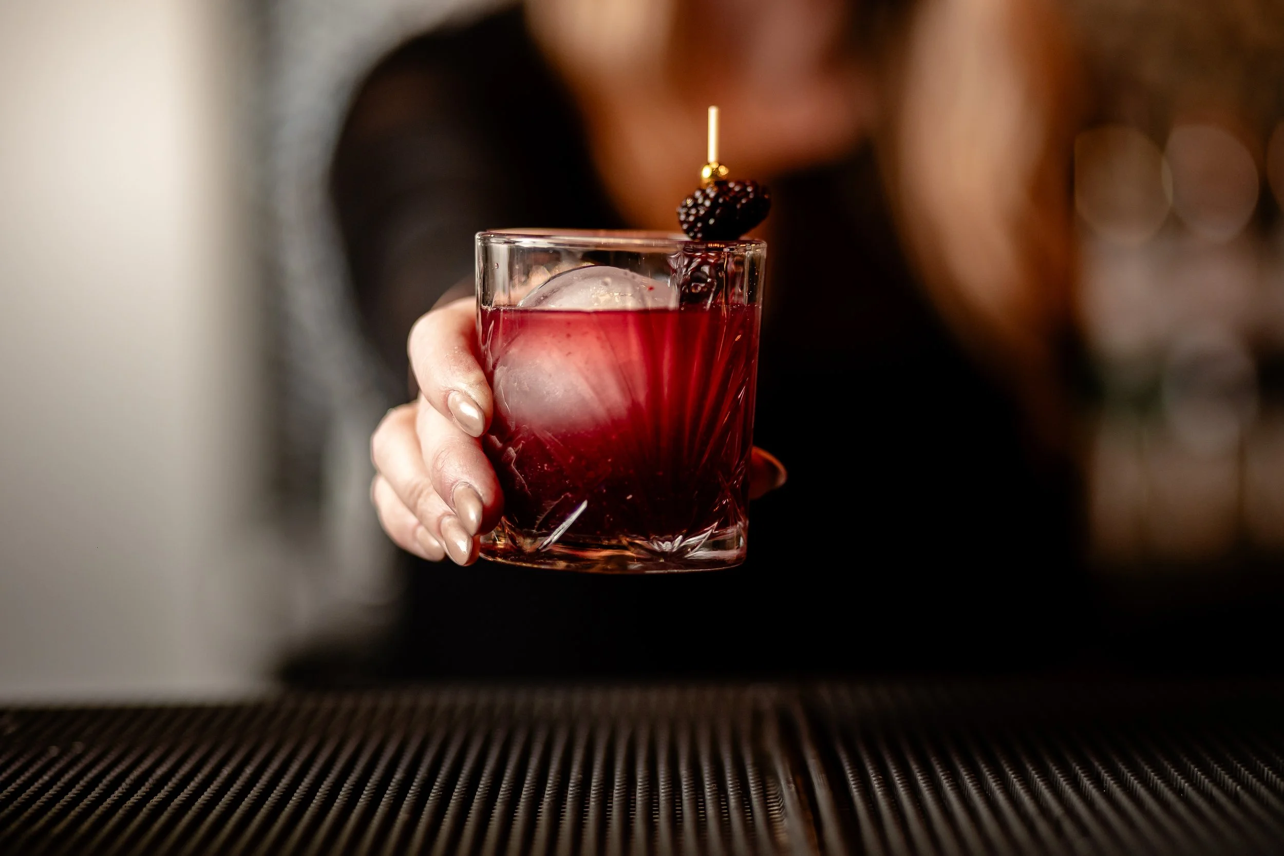 A hand holding a glass of red cocktail with an ice cube and a blackberry garnish, with a blurred background.