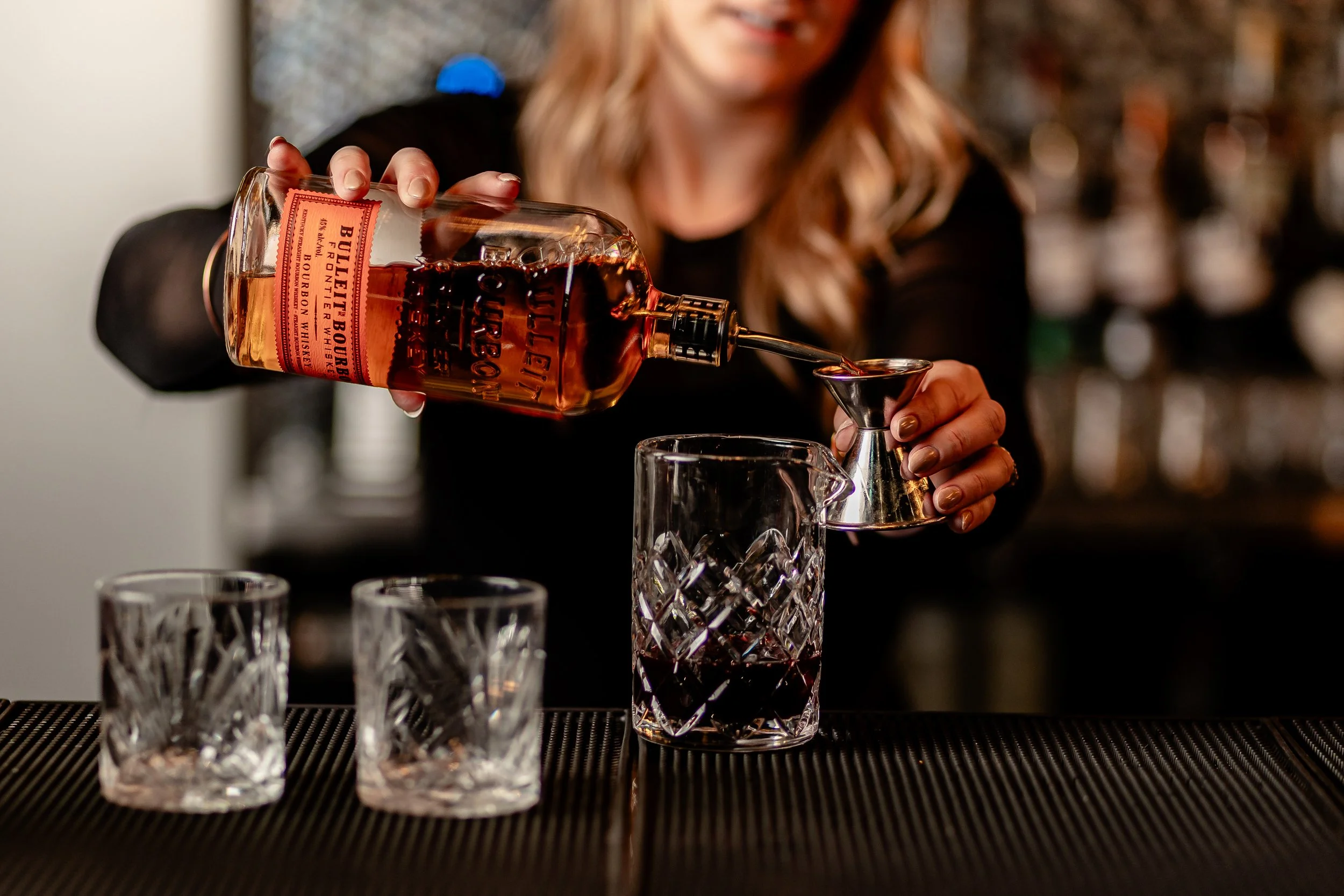 Person pouring whiskey into a glass using a jigger at a bar counter.