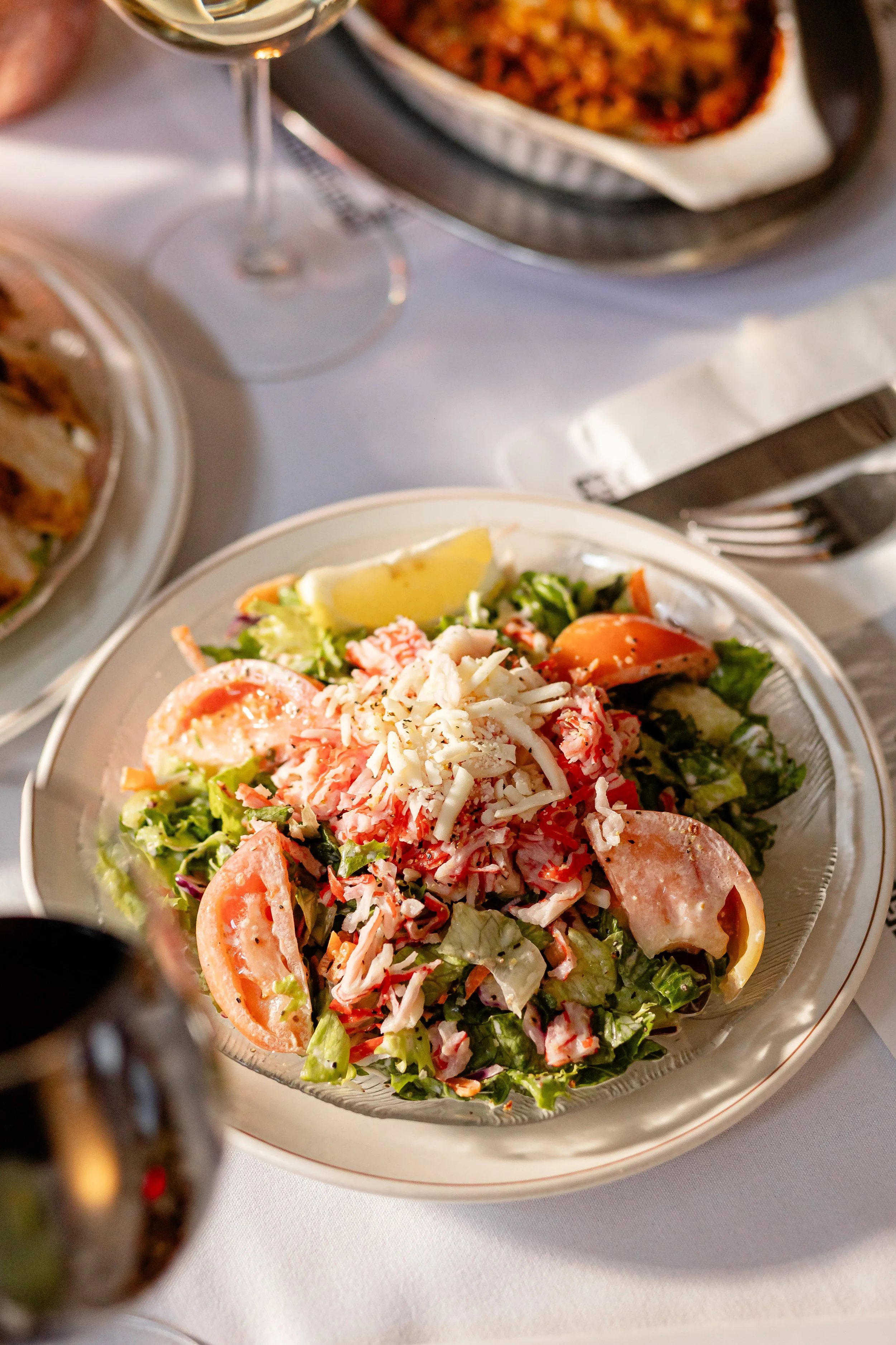 Close-up of a garden salad in a clear glass bowl, topped with shredded cheese and garnished with tomato slices and lemon wedges. Part of a tray with a baked dish and a glass of white wine are visible in the background.
