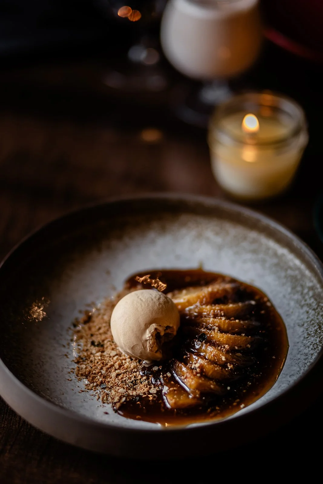 A dessert featuring sliced bananas drizzled with chocolate sauce, topped with a scoop of vanilla ice cream, and sprinkled with crushed nuts, served on a dark plate with a candle and a glass of drink in the background.