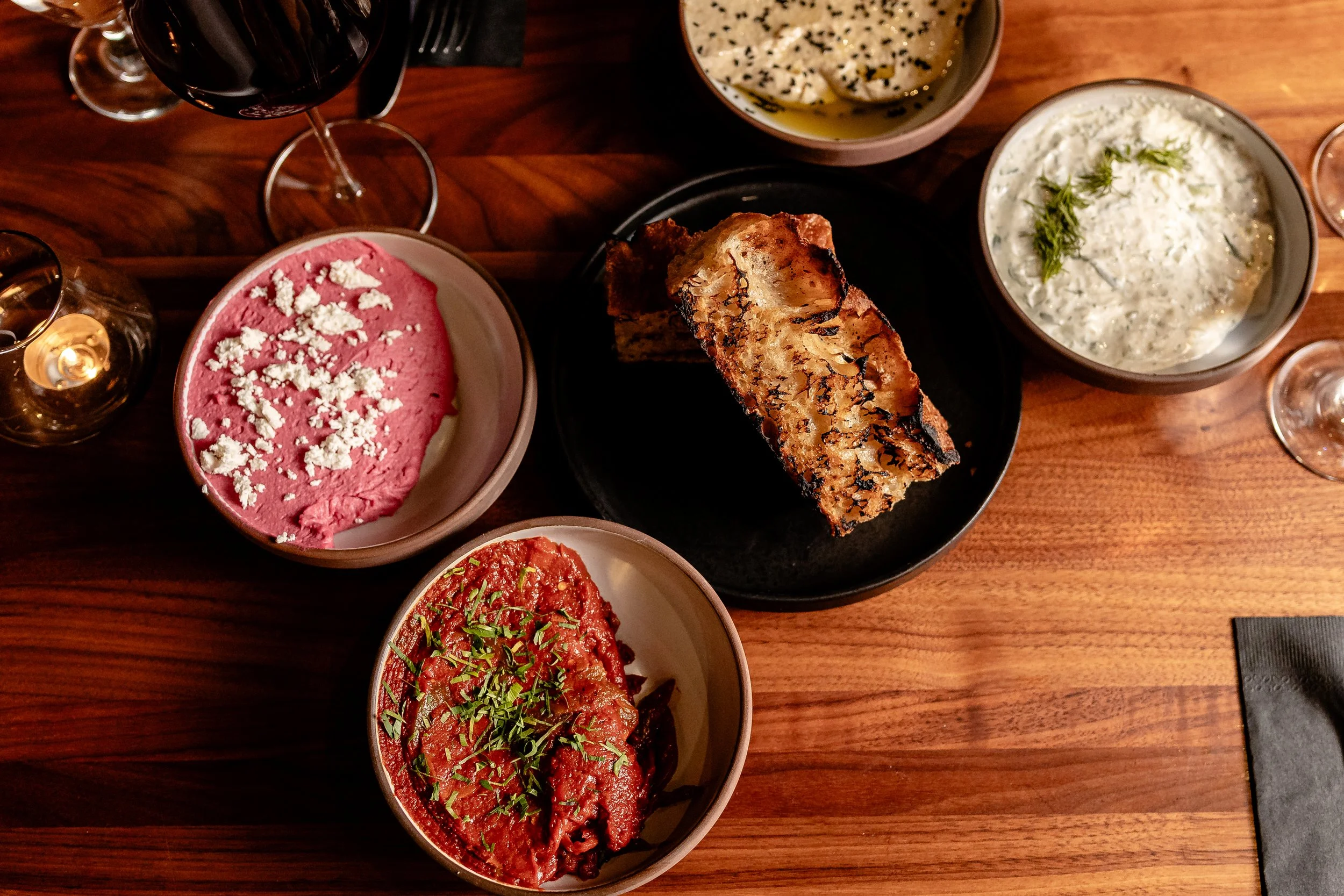 A top-down view of a dinner table with various bowls of food and a glass of red wine. The dishes include a bowl of pink hummus topped with crumbled feta, a bowl of creamy cucumber salad garnished with herbs, a bowl of tomato-based sauce with herbs, a