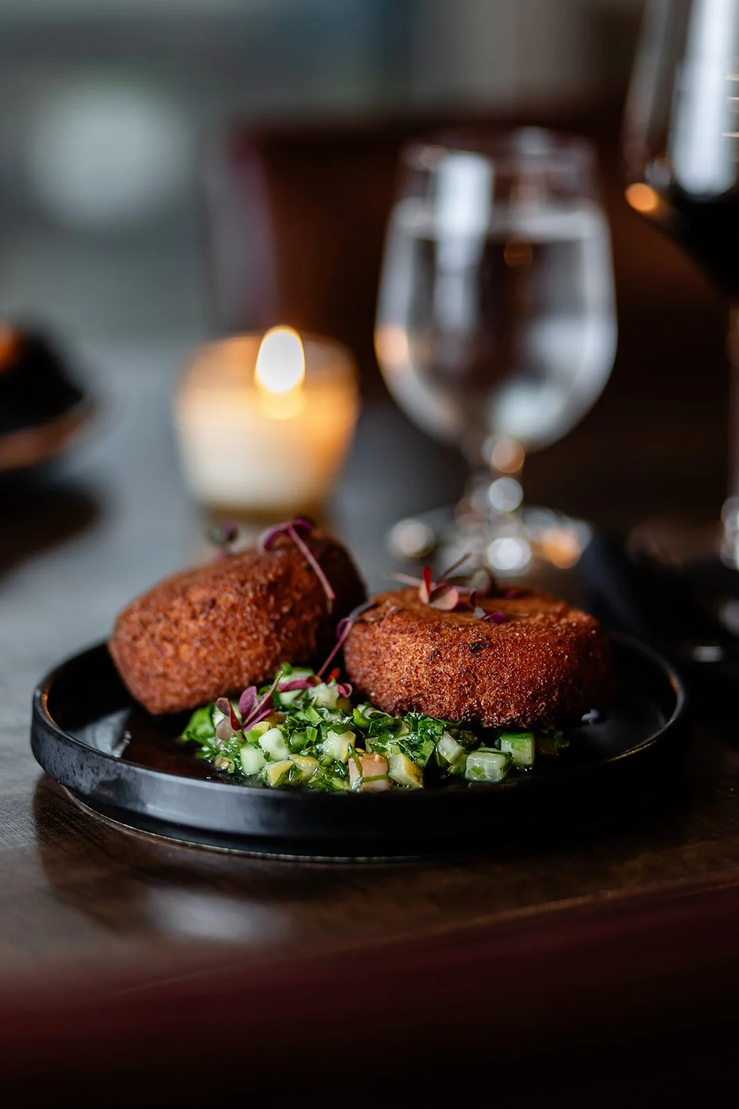 Fried croquettes on a black plate with a green cucumber salad and microgreens, glass of water, candle, and blurred wine glass in the background.