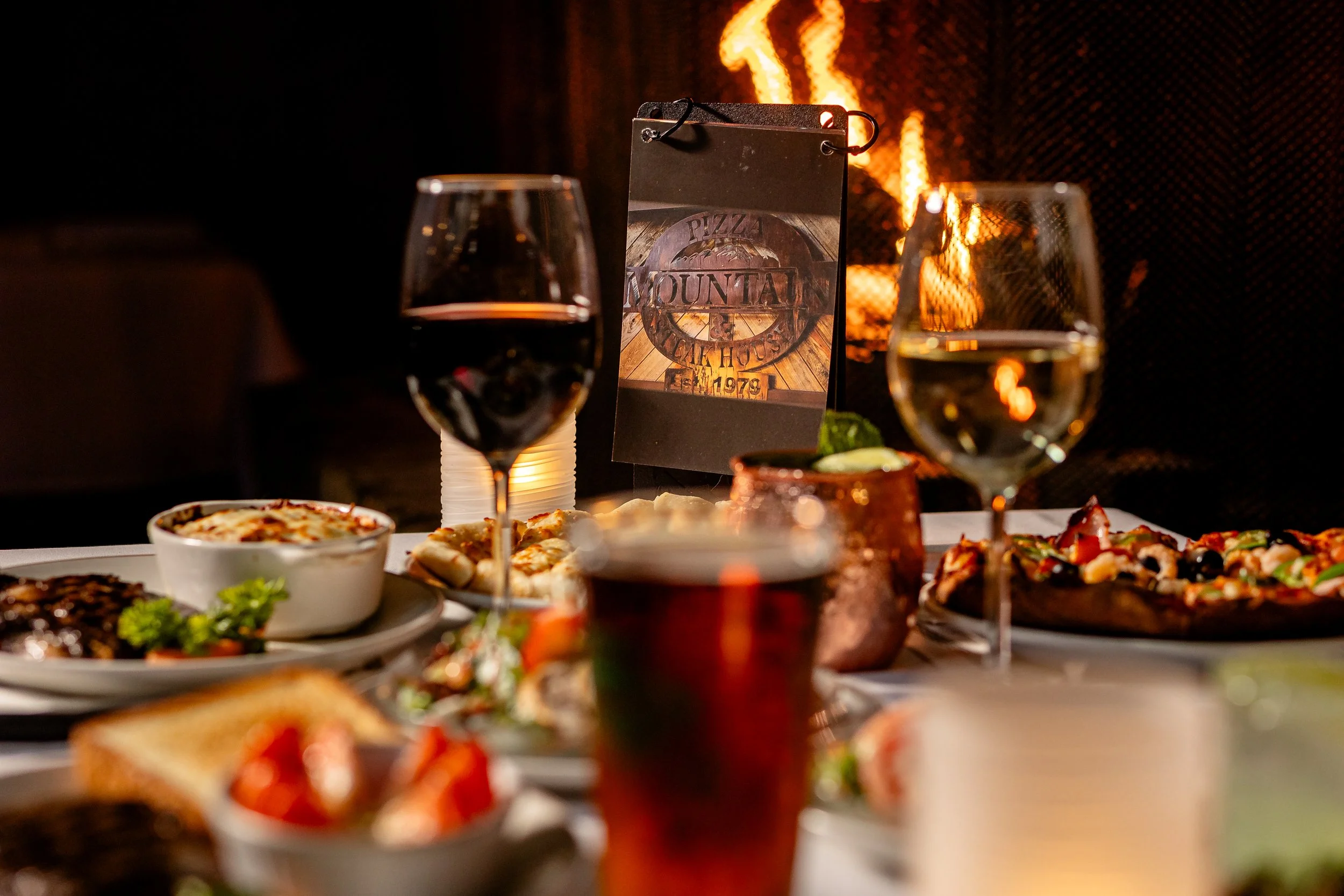 A table set with various dishes of pizza, salads, and drinks, with a pizza restaurant sign in the background and a fireplace creating a cozy atmosphere.