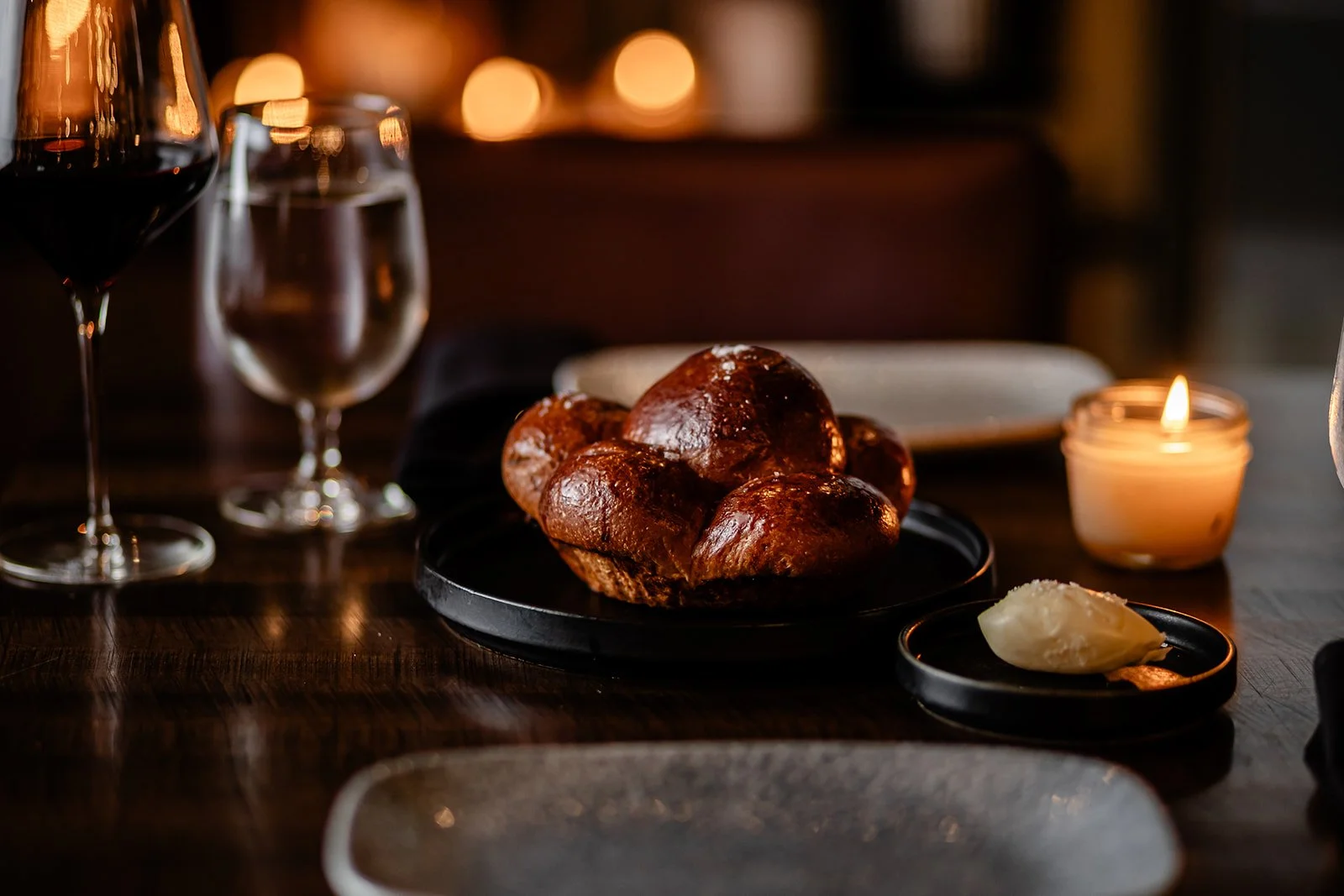 A cozy dinner setting with a plate of bread rolls, a dish of butter, a glass of red wine, a glass of water, and a lit candle on a wooden table.