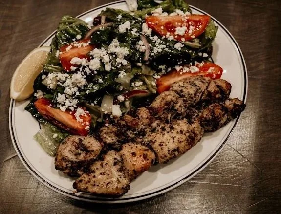 Plate of grilled meat with chopped salad topped with cheese, tomato, and lettuce, served with lemon wedge.
