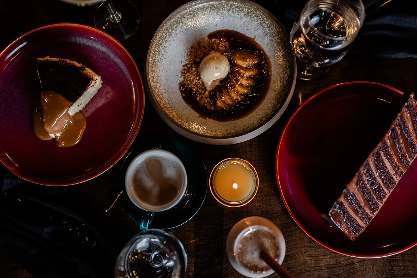 Desserts and drinks on a dark table, including a slice of cake with caramel sauce, a pudding with ice cream, another dessert with a striped pattern, a cup of coffee, a glass of water, and a wine glass.
