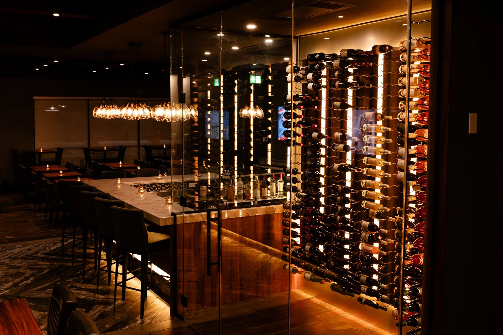 A dimly lit restaurant interior with a large wine rack filled with bottles on a glass wall, a marble-topped table with candles, and modern pendant lighting illuminating the space.
