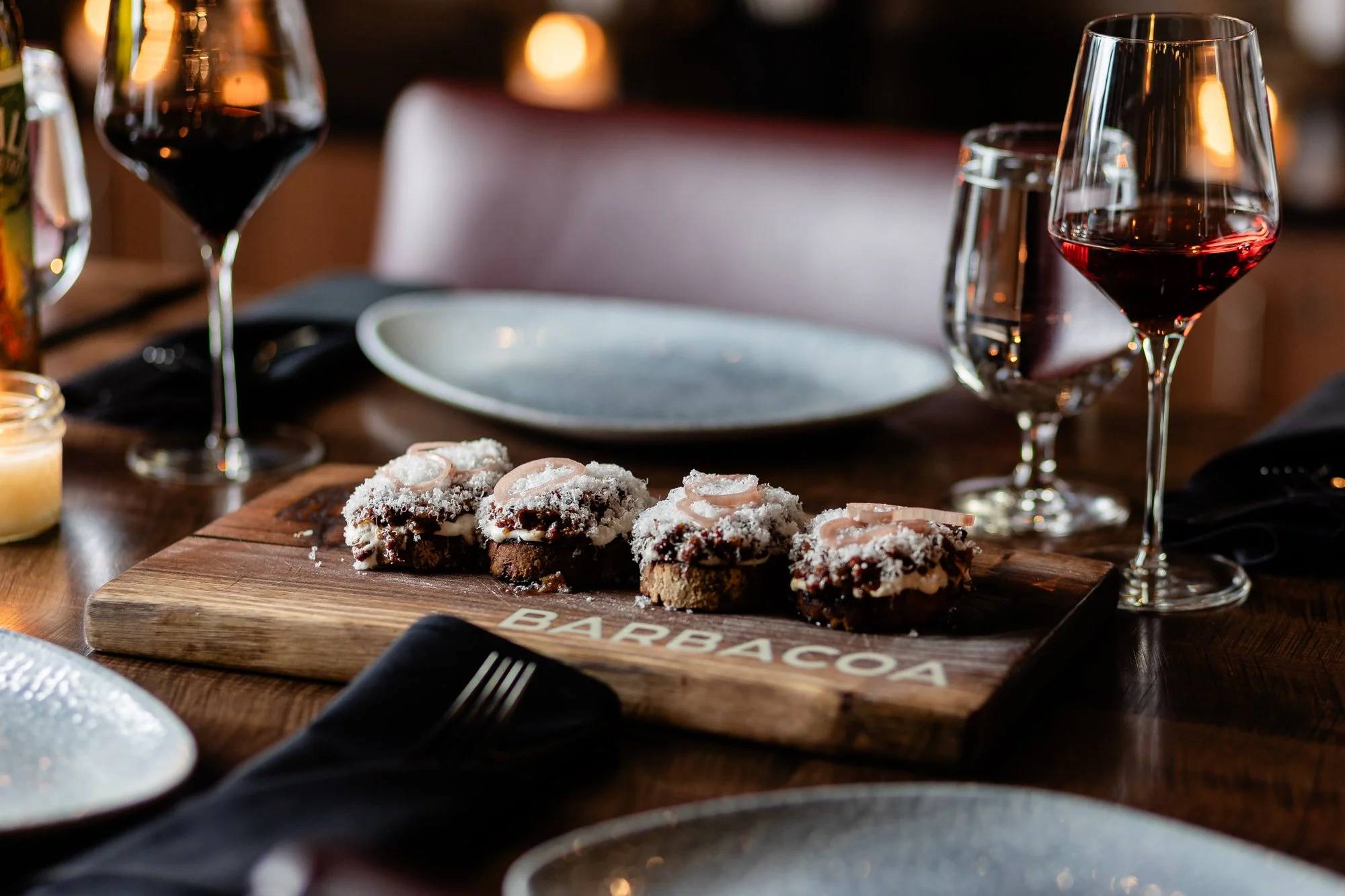 A wooden cutting board with several cupcakes topped with white frosting and sliced almonds. Behind the board are two glasses of red wine, a water glass, and a blurred background of a dining table.