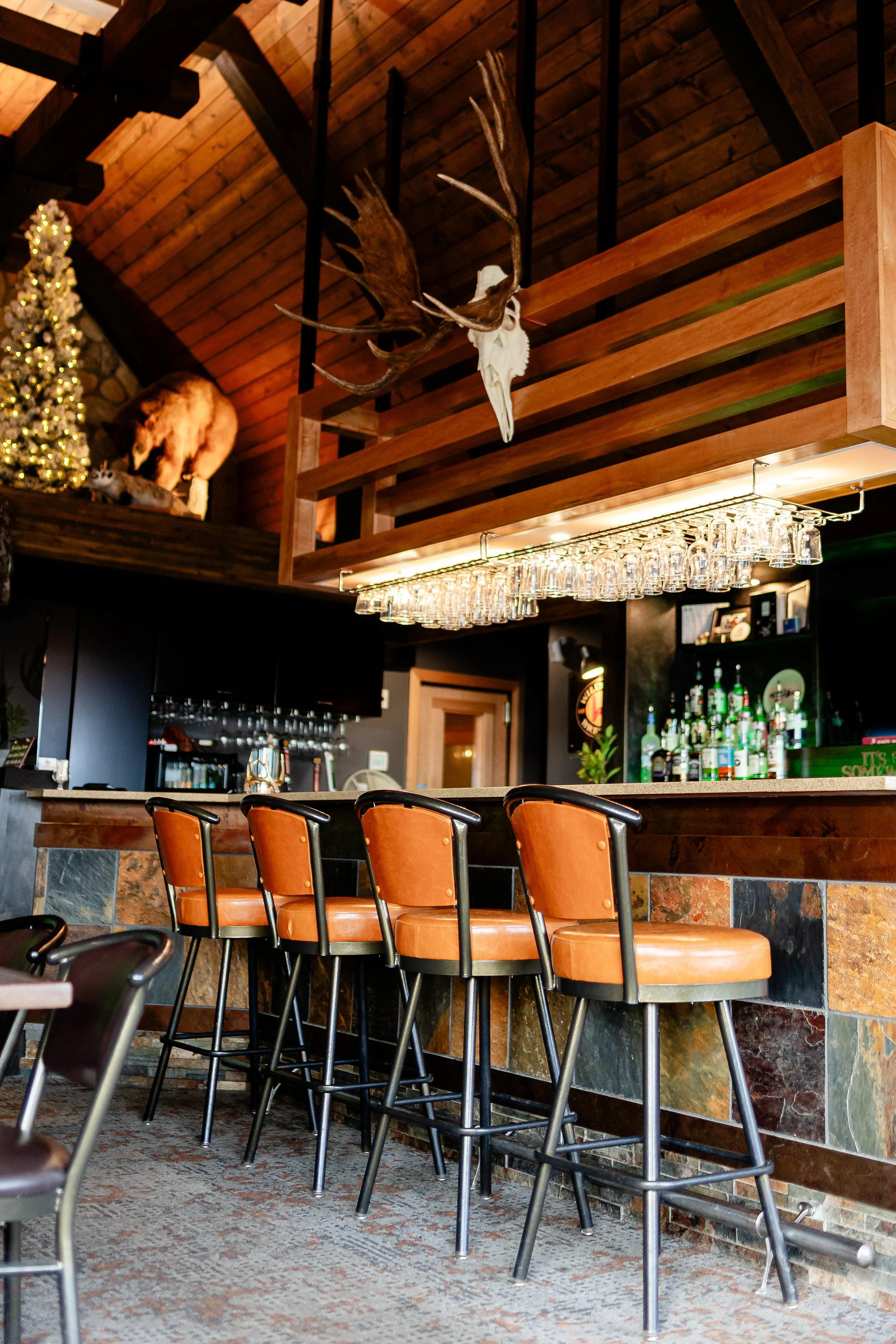 Interior of a rustic bar with wood accents, orange bar stools, a bar counter with liquor bottles, hanging glasses, decorative animal skull and antlers, and a Christmas tree
