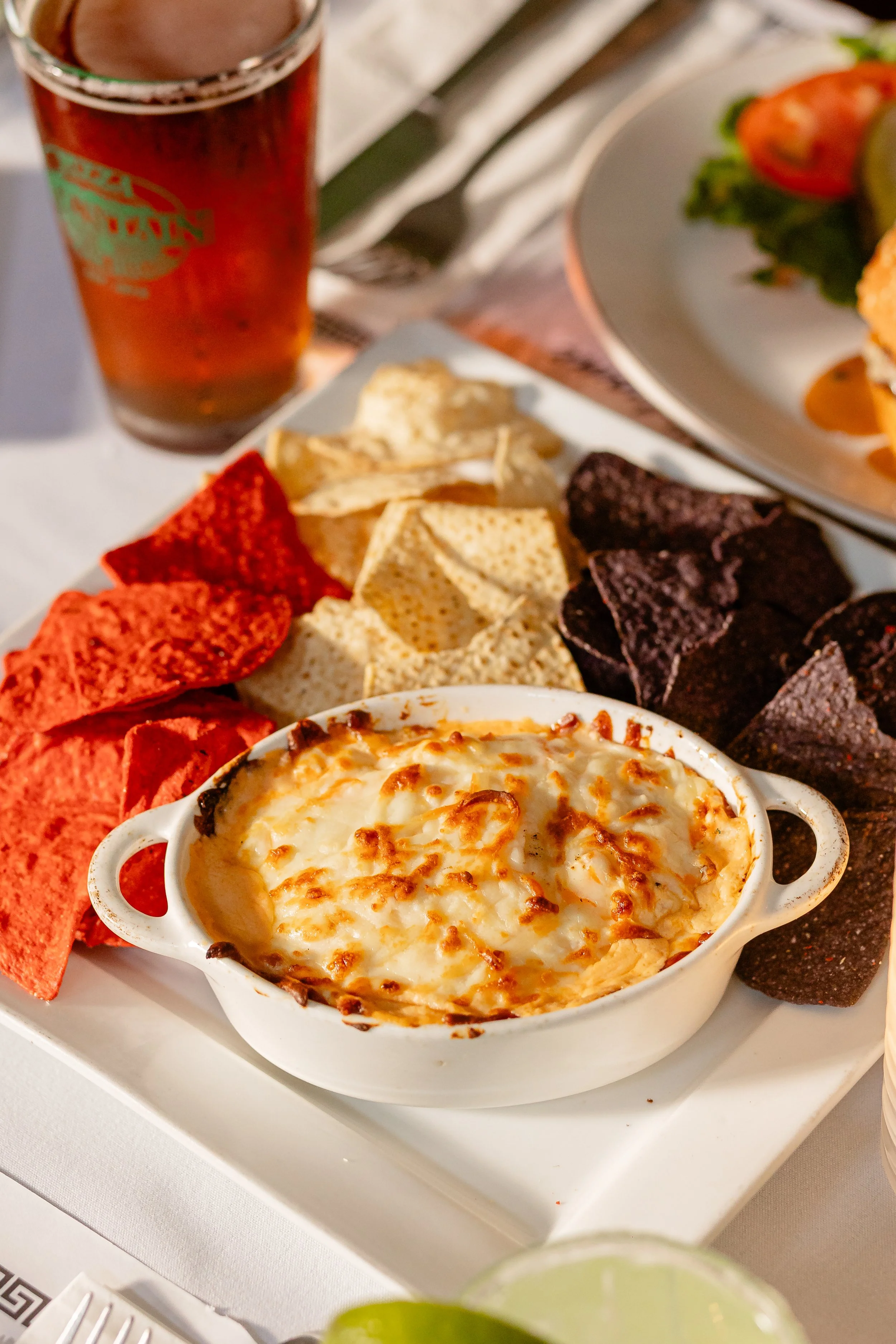 Baked cheesy dish in a small white bowl surrounded by red, white, and black potato chips, with a glass of a dark beverage in the background, on a white table.