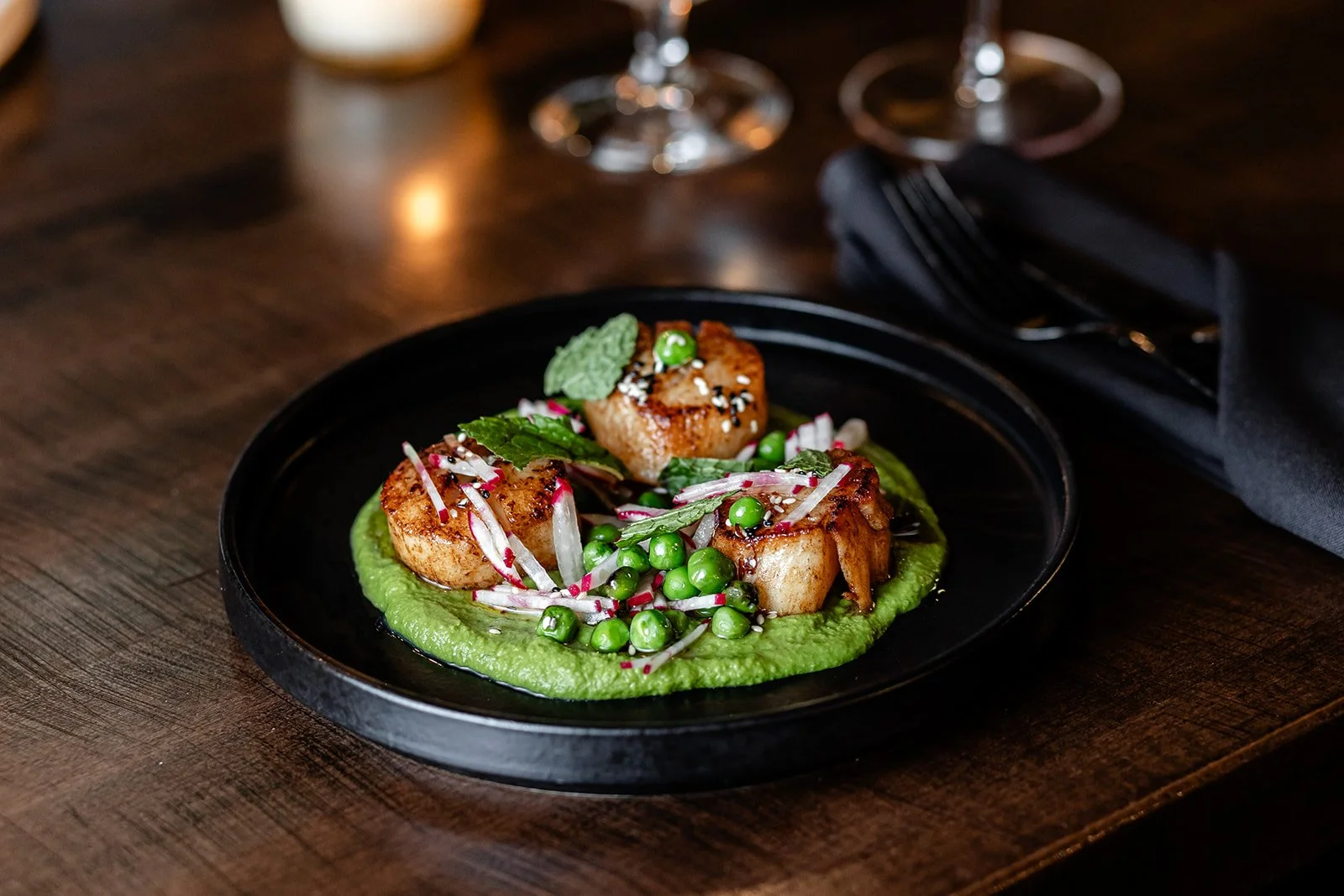 Green pea puree topped with seared scallops, microgreens, radish slices, and herbs, served on a black plate.