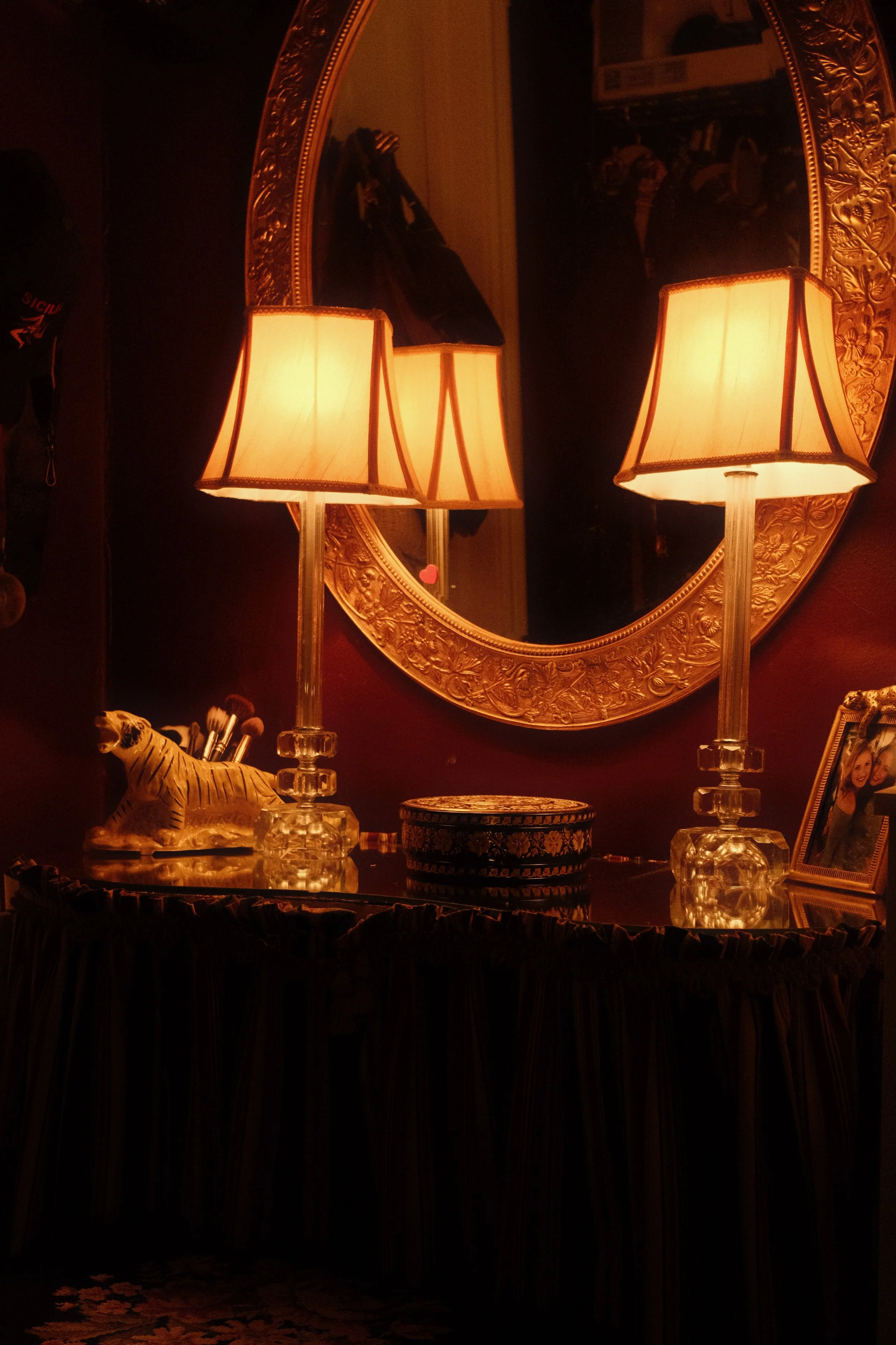 A decorative mirror with a gold ornate frame on a dark red wall, flanked by two glass table lamps with beige shades, a small tiger figurine, a decorative box, and a framed photo on a black table.