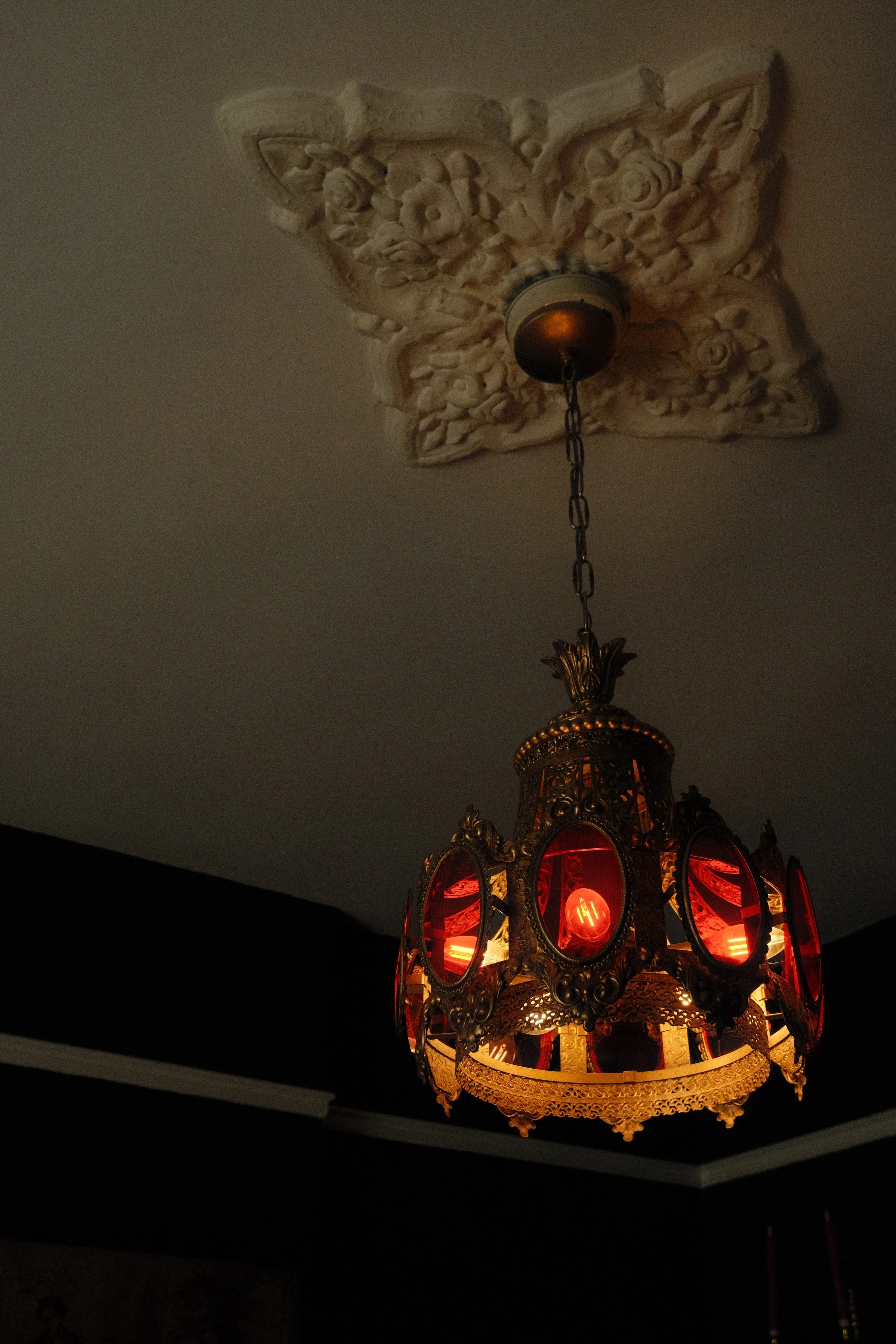A decorative hanging chandelier with intricate gold and red design and illuminated red lights, hanging from an ornate ceiling medallion in a dark room.