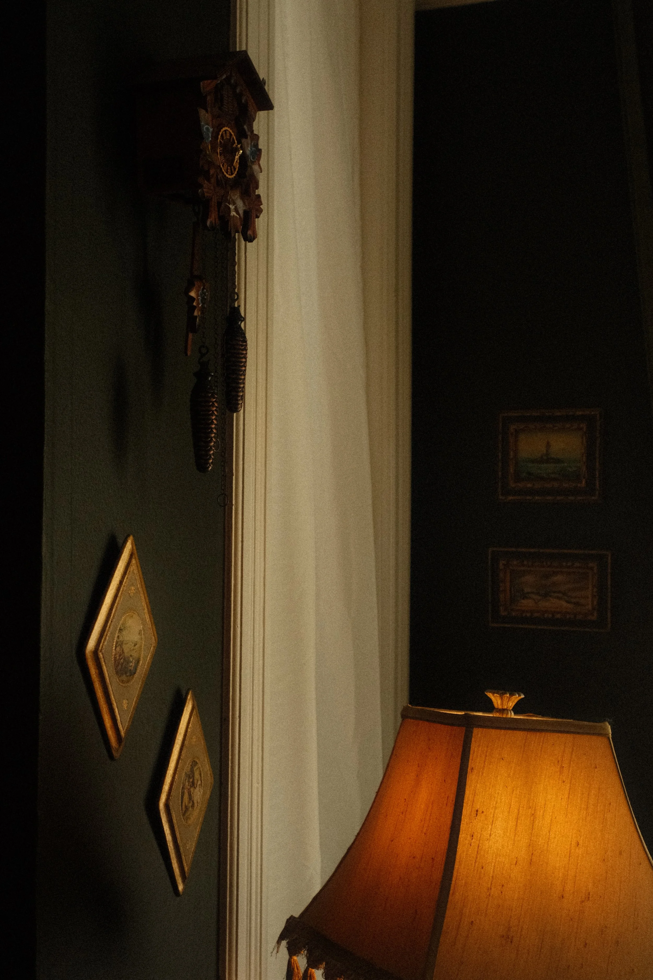 A dimly lit room with a dark-colored wall decorated with framed artwork. A wooden table with a yellow lampshade sits in the foreground. Along the wall, there is a cuckoo clock and several framed pictures hanging.