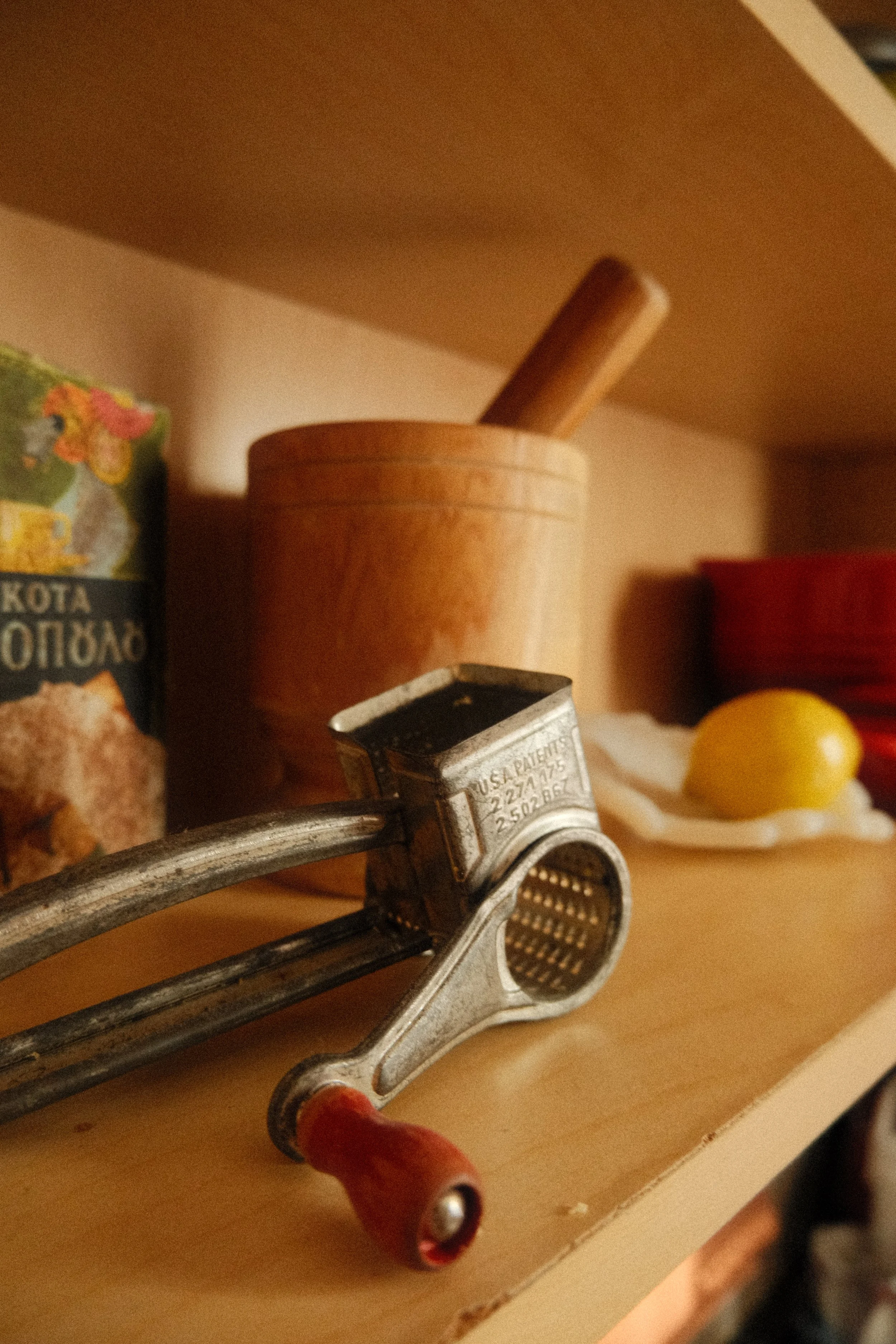Close-up of a vintage garlic press with a red handle, resting on a wooden surface. In the background, there is a small container or jar and a yellow object, possibly a lemon, blurred out.