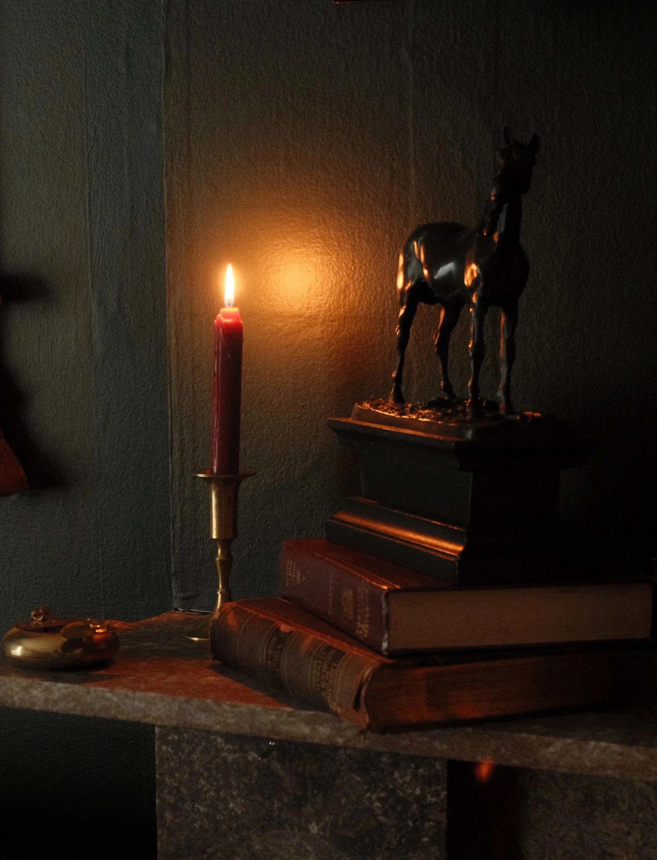 A dark room illuminated by a single lit red candle, a bronze horse sculpture on a shelf, and a stack of books with one open at the bottom.