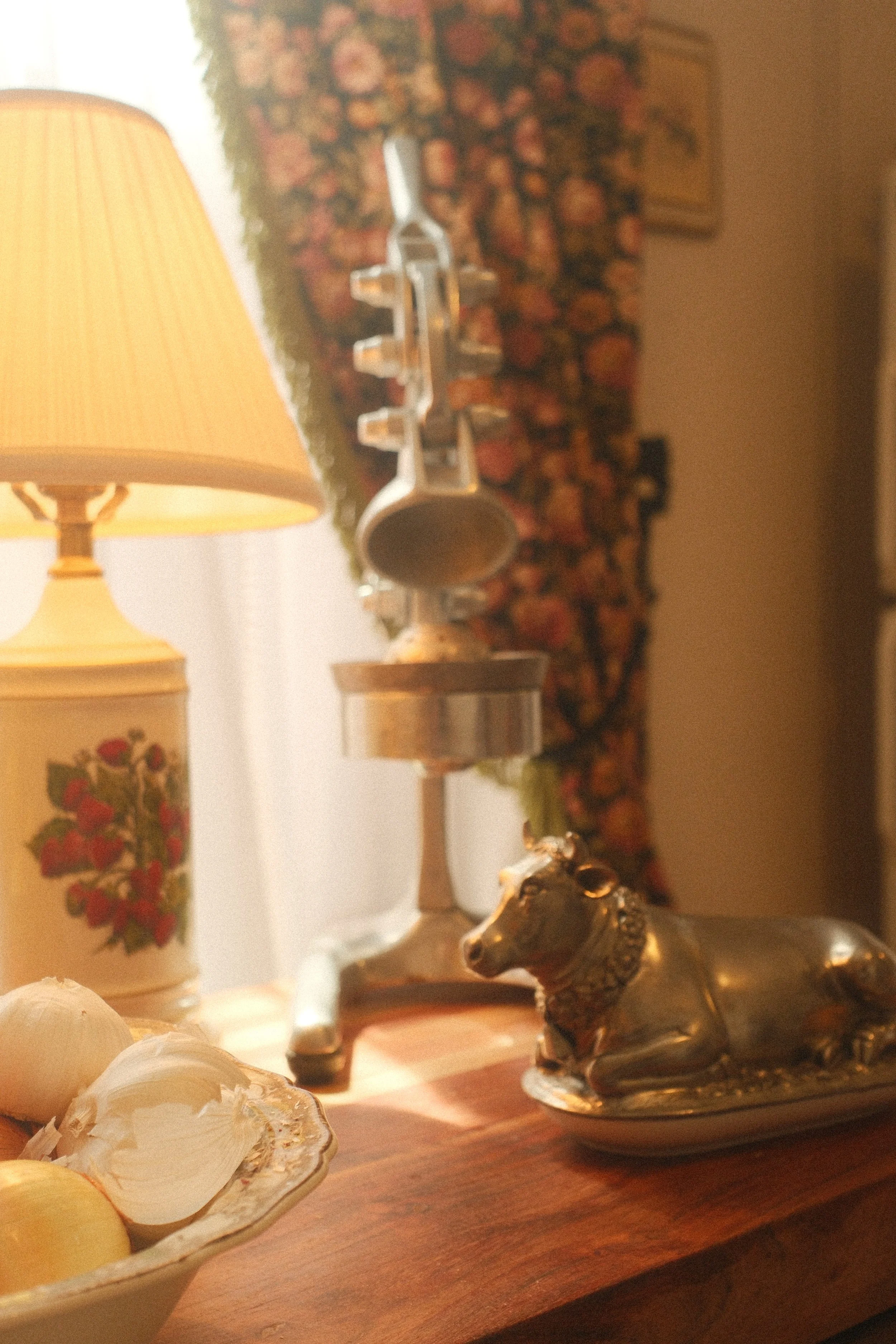 A collection of decorative items on a wooden surface, including a vintage lamp with a floral design, a brass elephant figurine, and other tableware. The background features a floral-patterned fabric or curtain and a framed picture on the wall, giving a cozy, vintage interior ambiance.