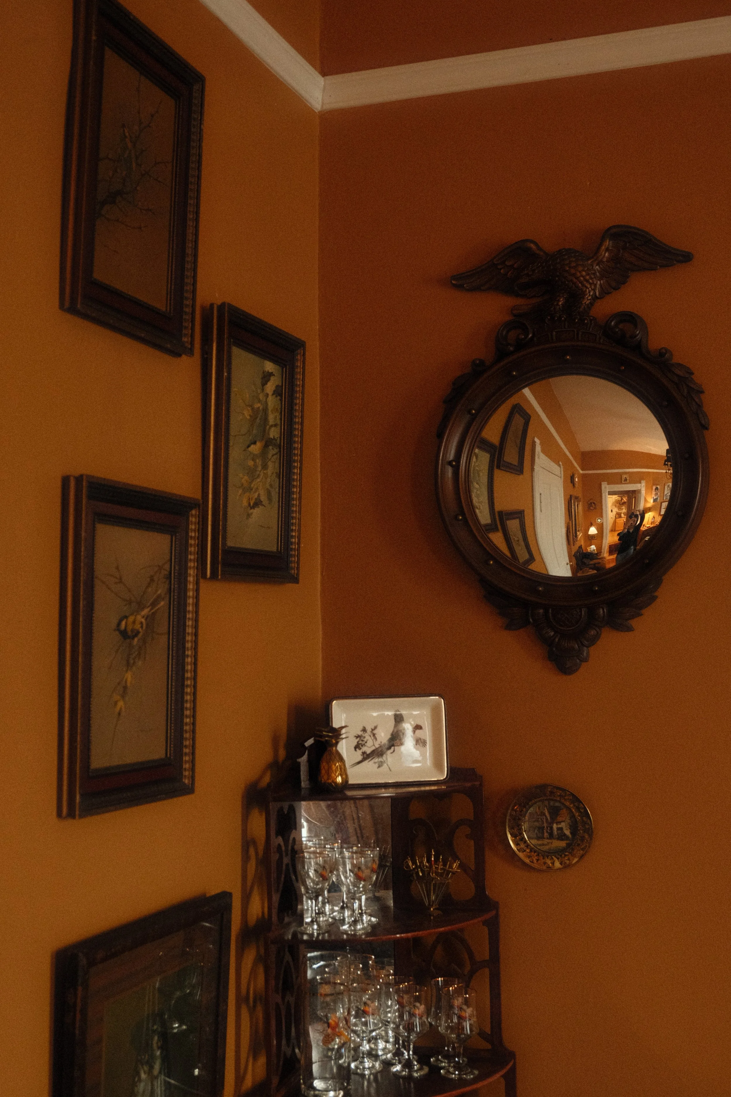 A corner of a room with warm orange walls decorated with framed pictures of flowers and nature, a wall mirror with a carved eagle on top, and a wooden cabinet filled with glassware and decorative items.