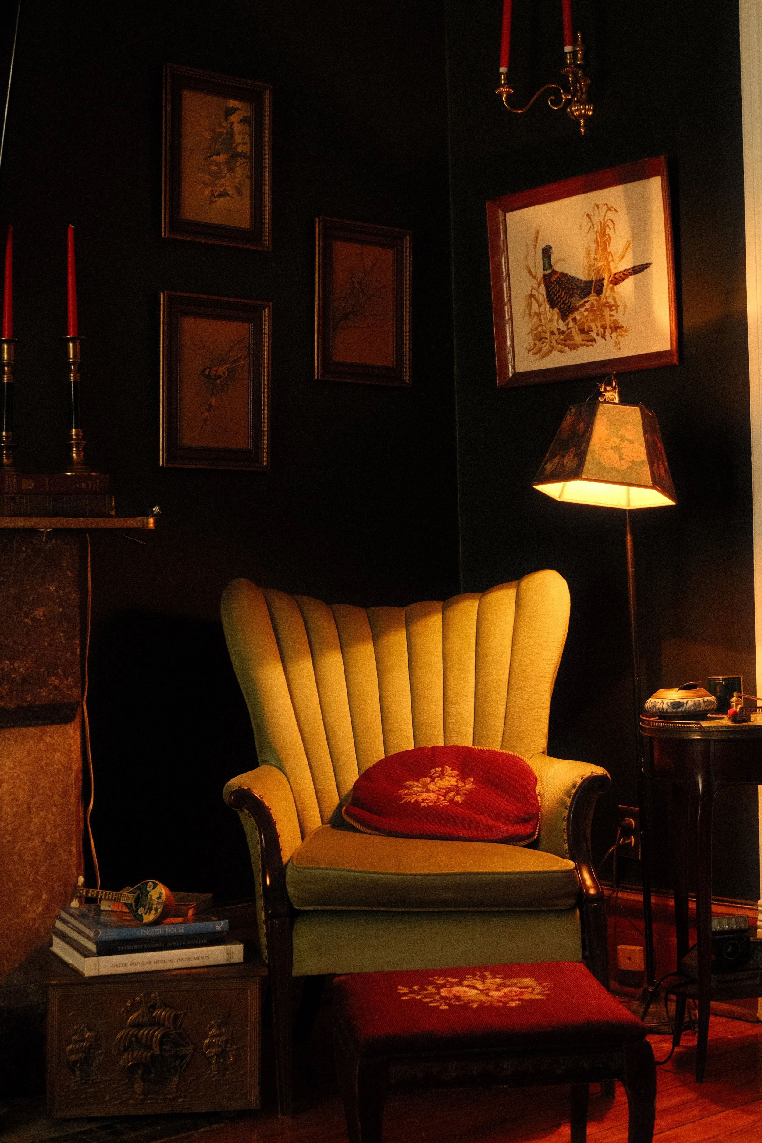 A cozy living room corner with a vintage green armchair with a red pillow in front of a dark wall. To the right, there's a small black side table with decorative items and a lamp with a floral shade. The dark wall is decorated with framed artwork depicting birds and nature, illuminated softly by warm lighting from a chandelier and lamp.
