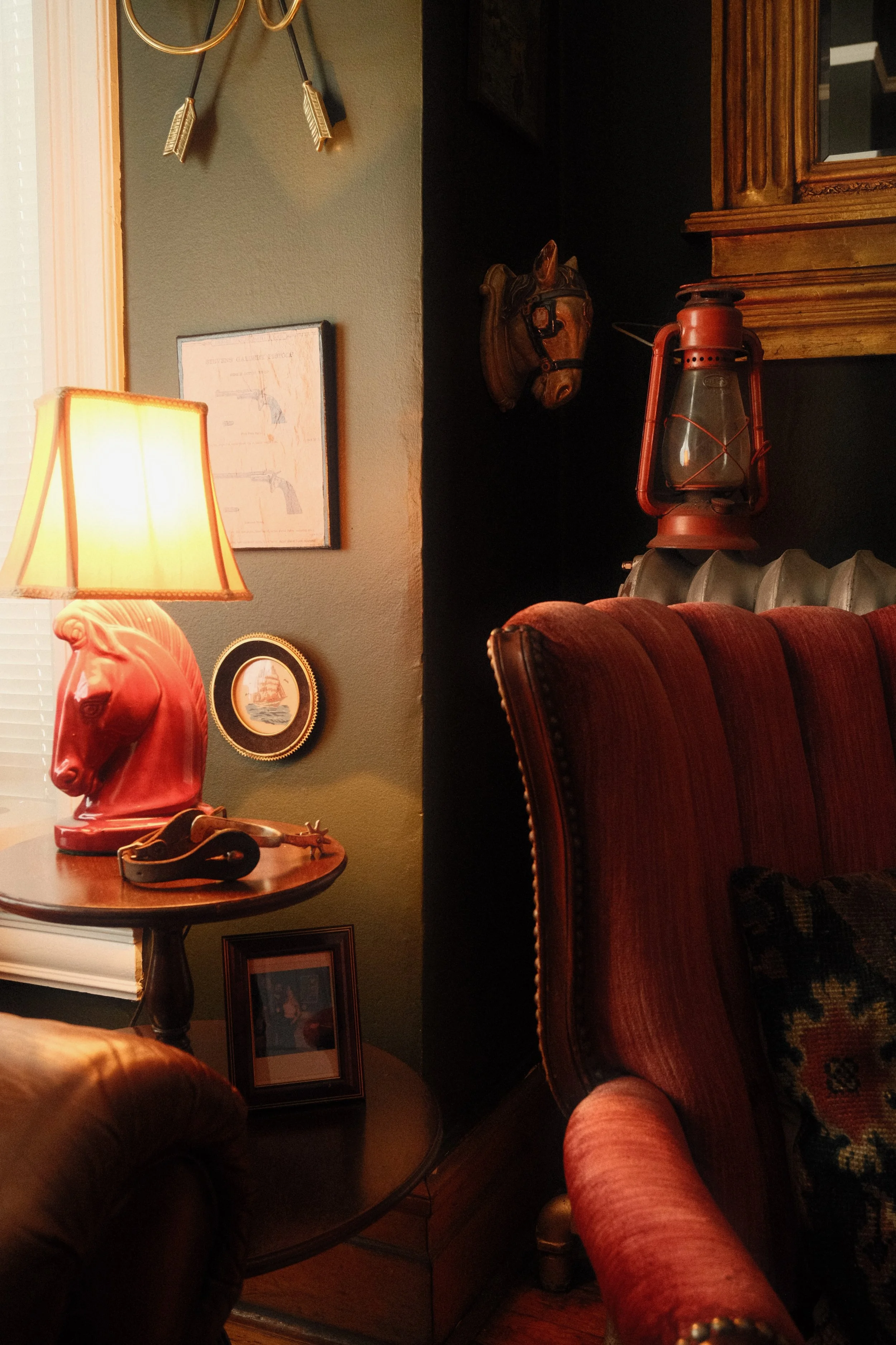 A cozy living room with a vintage theme, featuring a red cow-shaped lamp, a wooden side table with a pair of glasses and a decorative item, and framed pictures on the wall. There is also a mounted horse head and an antique lantern on a shelf, with a plush armchair in the foreground.
