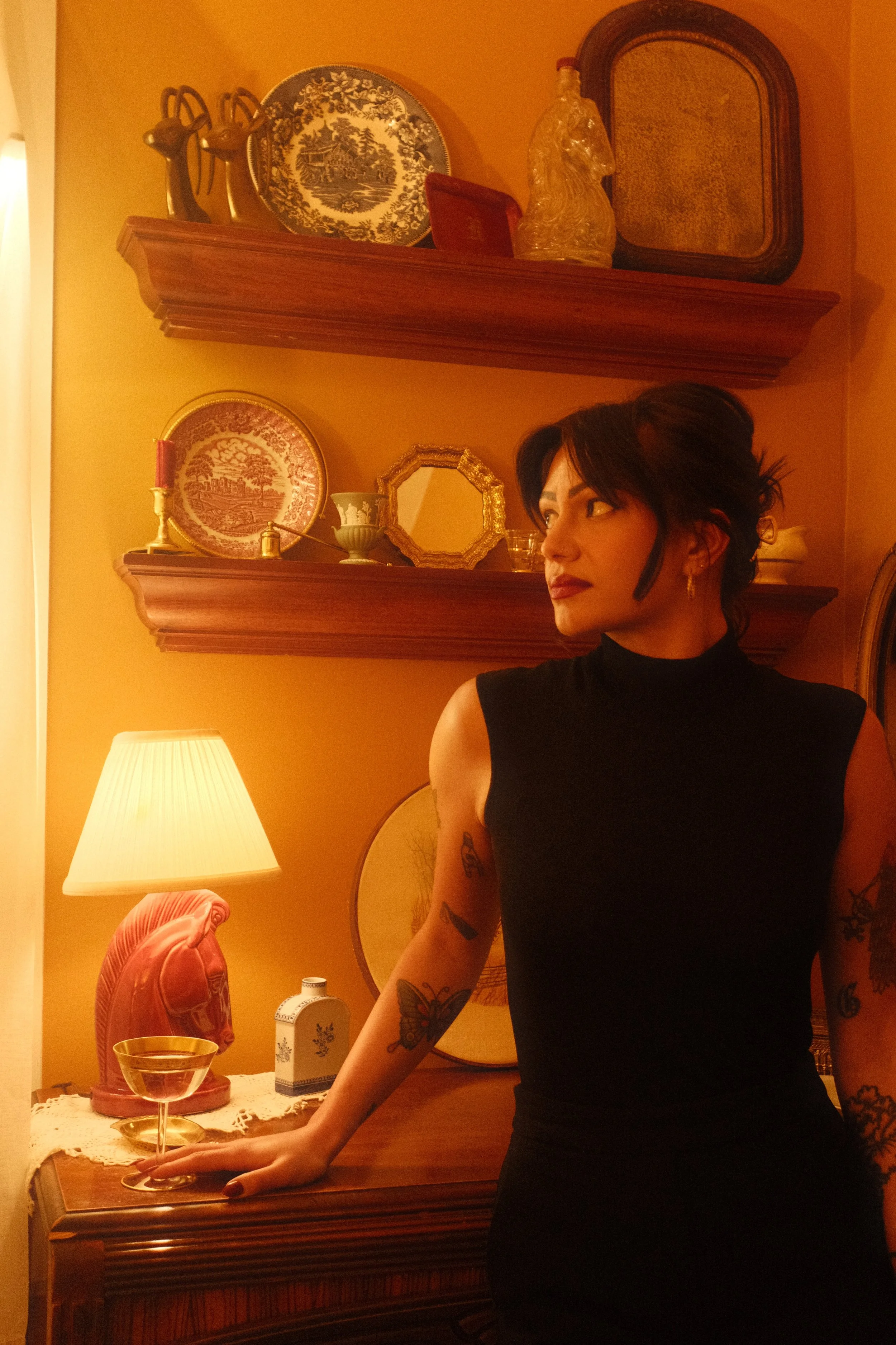 A woman with dark hair and tattoos on her arms, wearing a black sleeveless top, standing in a warmly lit room with antique decor and shelves with decorative plates and items.