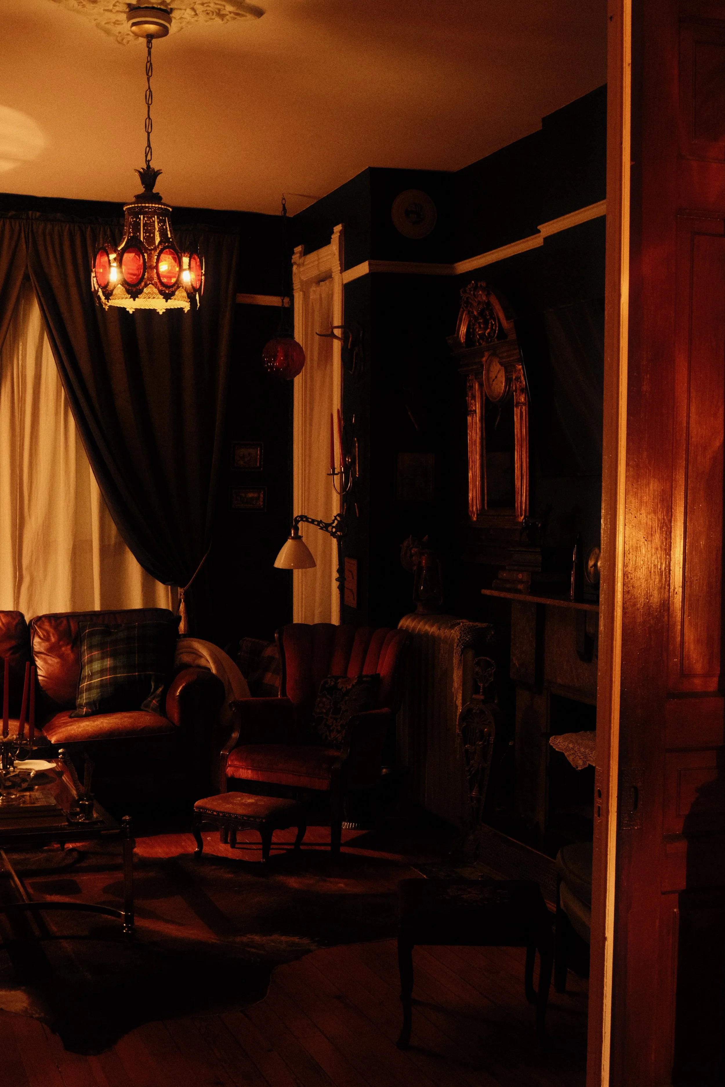 A cozy, dimly lit living room with vintage furniture, dark curtains, a chandelier with red accents, an ornate clock on the wall, and various decorative items.