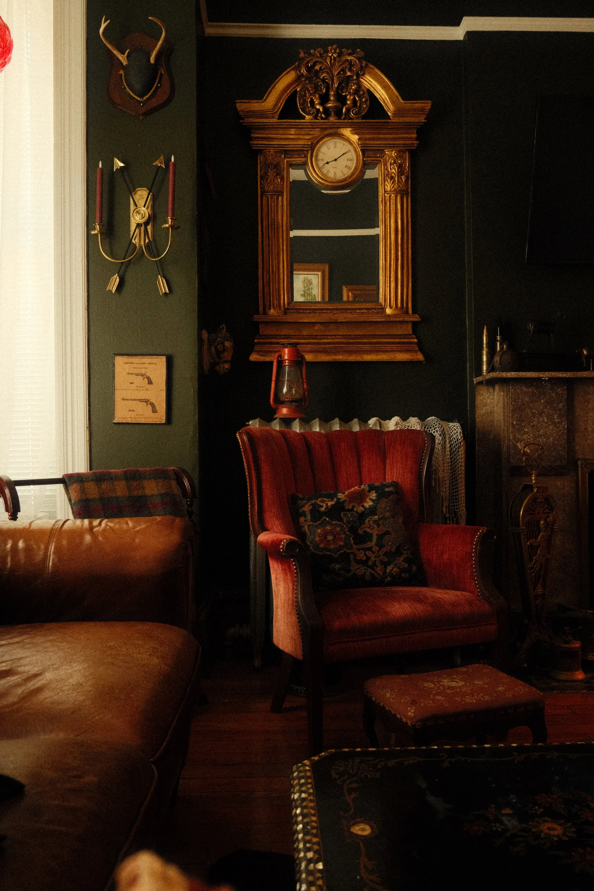 Vintage wooden wall clock with a round face and Roman numerals, mounted on a dark wall with ornate carvings at the top. To the left, a green wall with a mounted deer head and a wall sconce with two candles. Several upholstered chairs with patterned fabric in a dimly lit room.