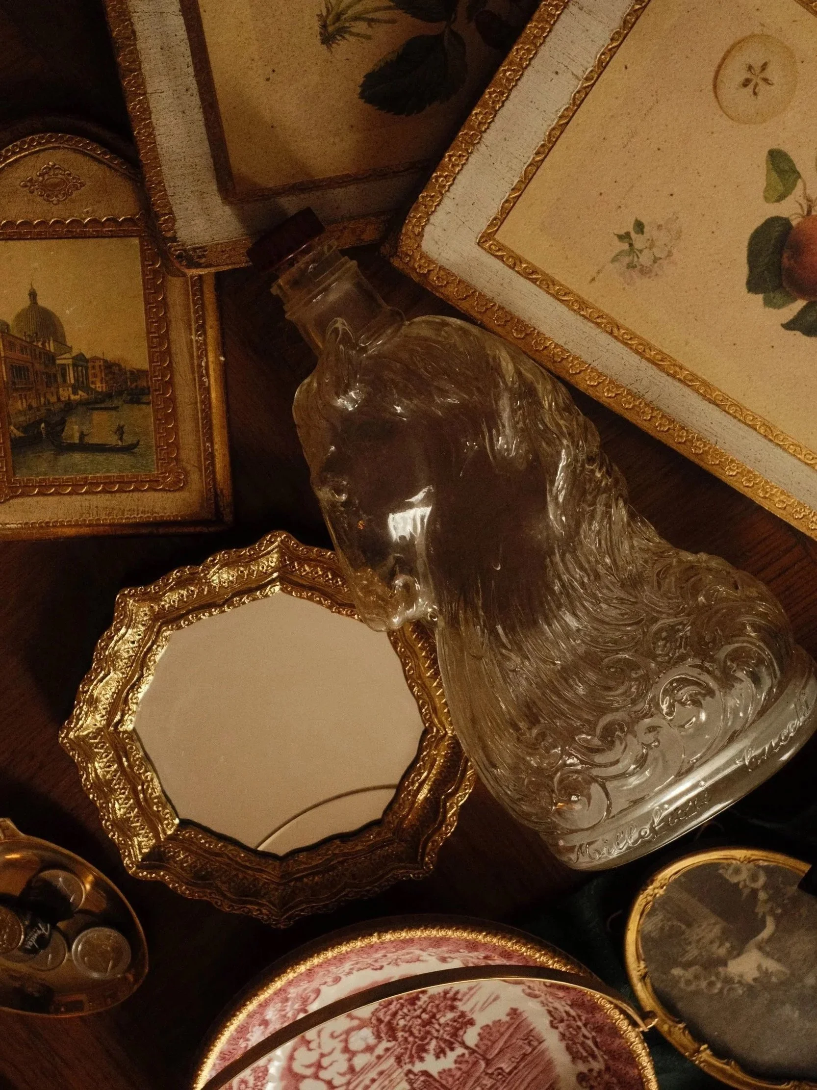 A collection of decorative trays and a clear glass decanter with a dark cap on a wooden surface. The trays have ornate gold edges and vintage-style artwork depicting scenes and floral designs.