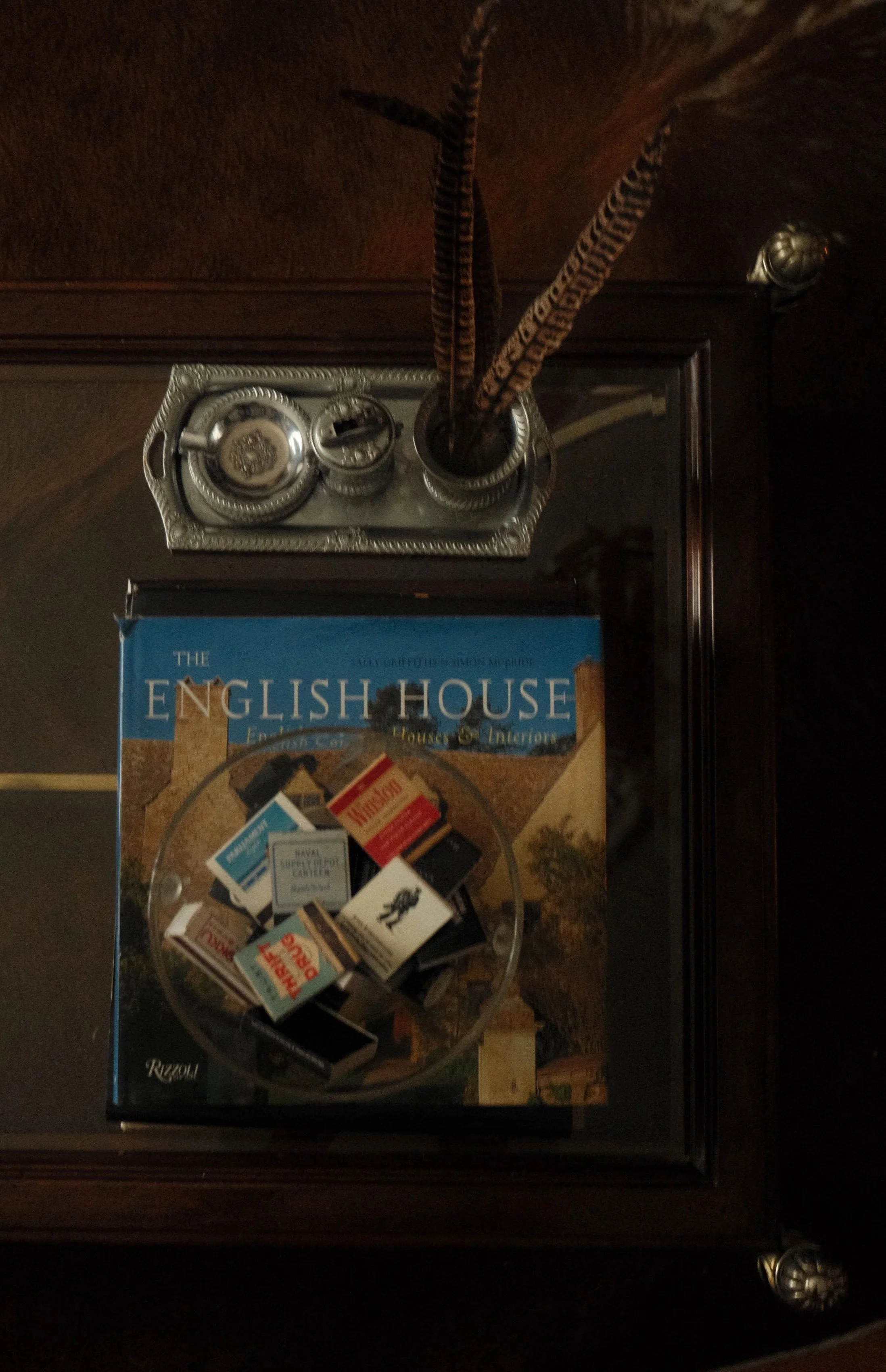 Top of a wooden cabinet showing a silver-colored tray with closed small containers, a book titled 'The English House,' and a clear glass bowl filled with matches and small packets of matches.