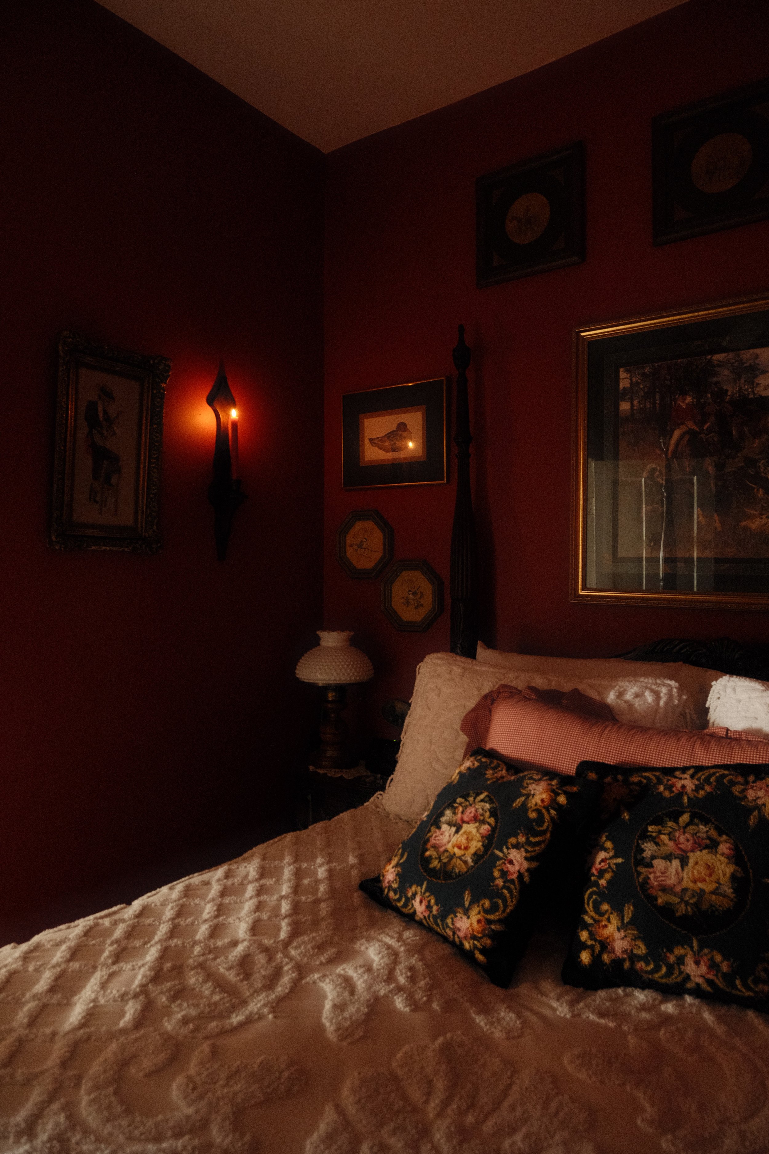 A cozy bedroom with dark red walls decorated with framed pictures and paintings. A bedside table with a vintage-style lamp and a pillow with a floral design resting on a bed with a textured, scalloped bedspread.