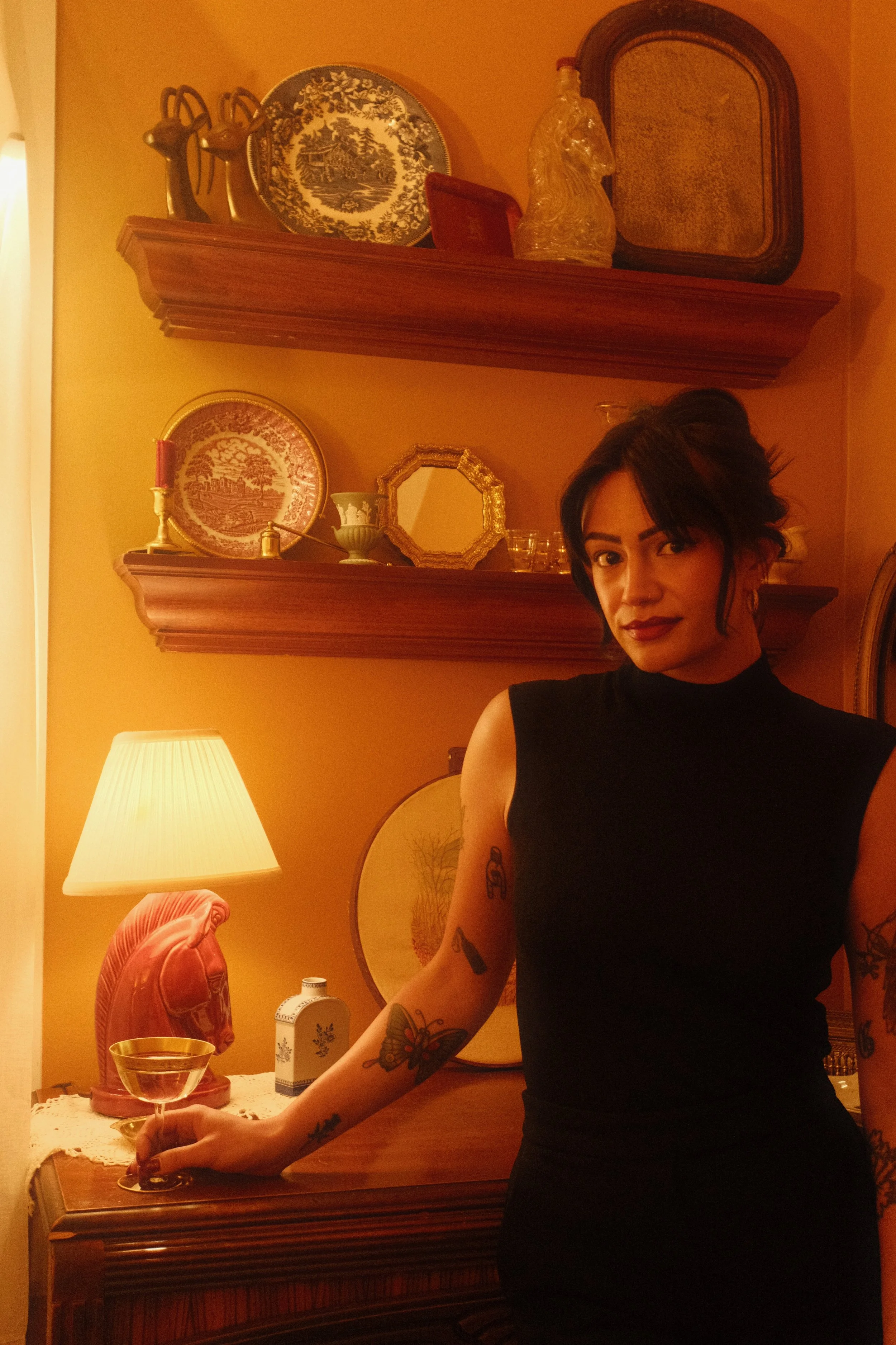A woman with dark hair styled in a messy bun, wearing a sleeveless black top, standing next to a wooden sideboard in a warmly lit room. She has tattoos on her left arm, including a butterfly on her forearm and a small design on her upper arm, and is holding a glass of champagne. Behind her, there are vintage decorative items on the sideboard, including a pink ceramic sculpture, a white bottle with floral design, and a lamp with a pleated shade. The yellow walls are decorated with ornate plates, framed artwork, and wooden shelves displaying decorative objects and ceramics.