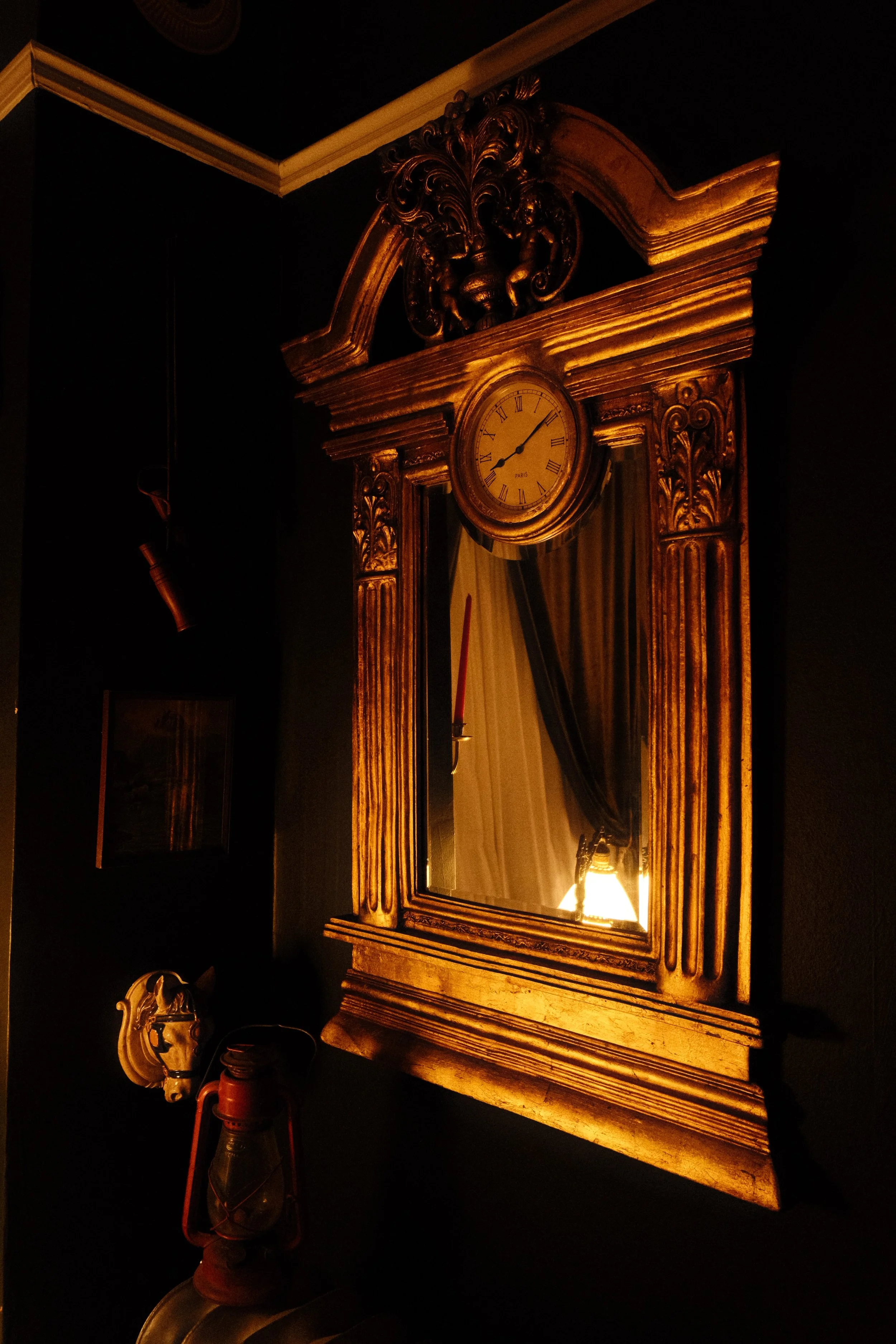 A gold ornate wall clock with Roman numerals, mounted above a mirror. The clock shows the time as 12:07. The mirror reflects part of the room. The surrounding decor includes a small skull and a vintage lantern on a table below.