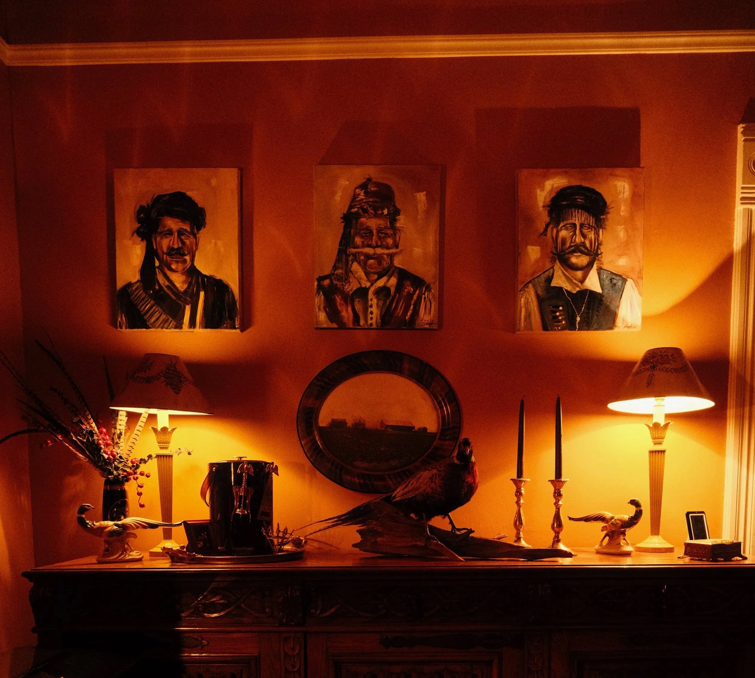 Decorative wall with three framed portraits, lamps, and various ornamental items on a darkwood table in a warmly lit room.