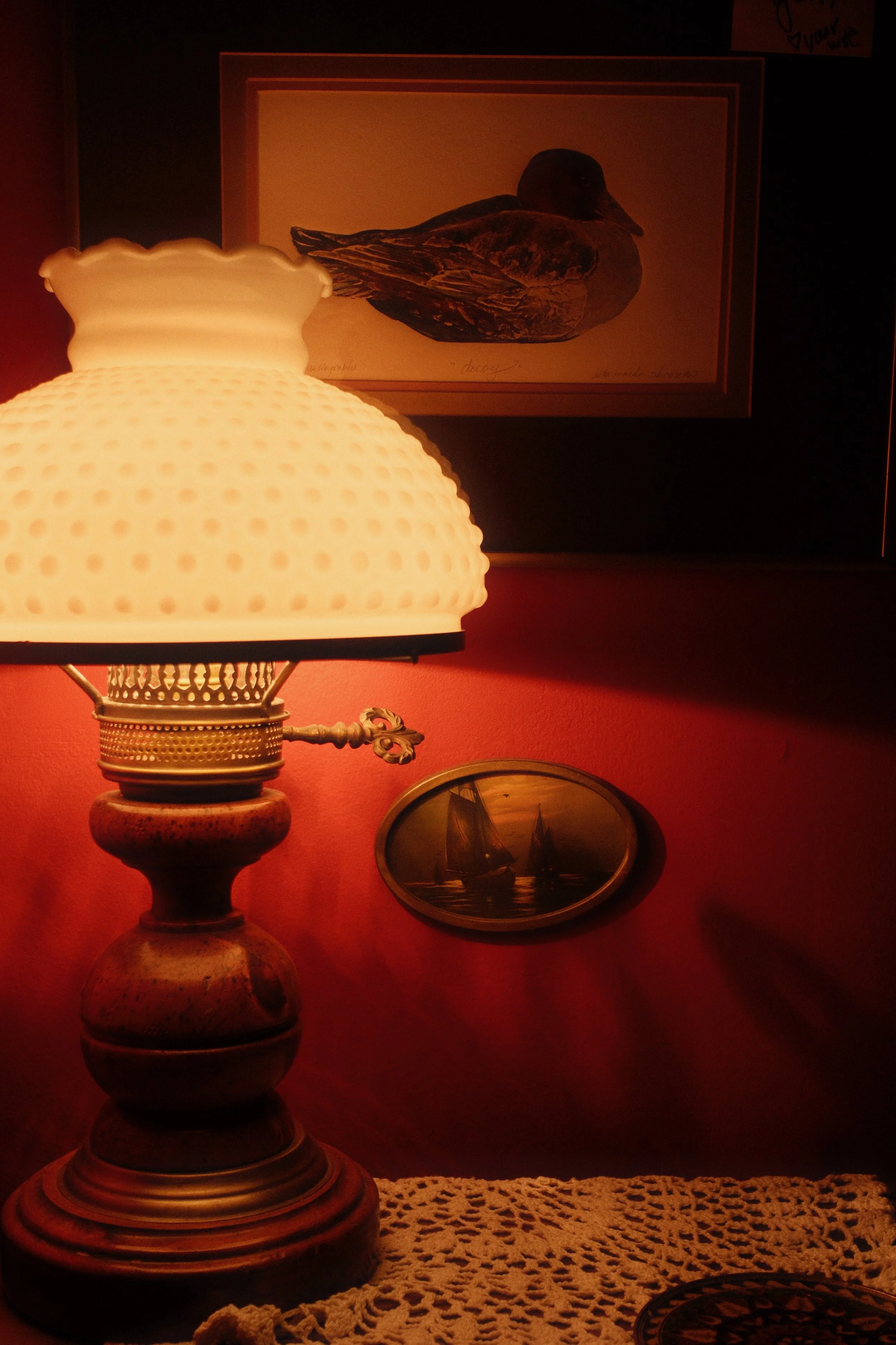 A vintage table lamp with a textured white glass shade, placed on a crocheted tablecloth. In the background, there is a framed illustration of a duck or waterfowl, and an oval painting of sailboats on water on a red wall.