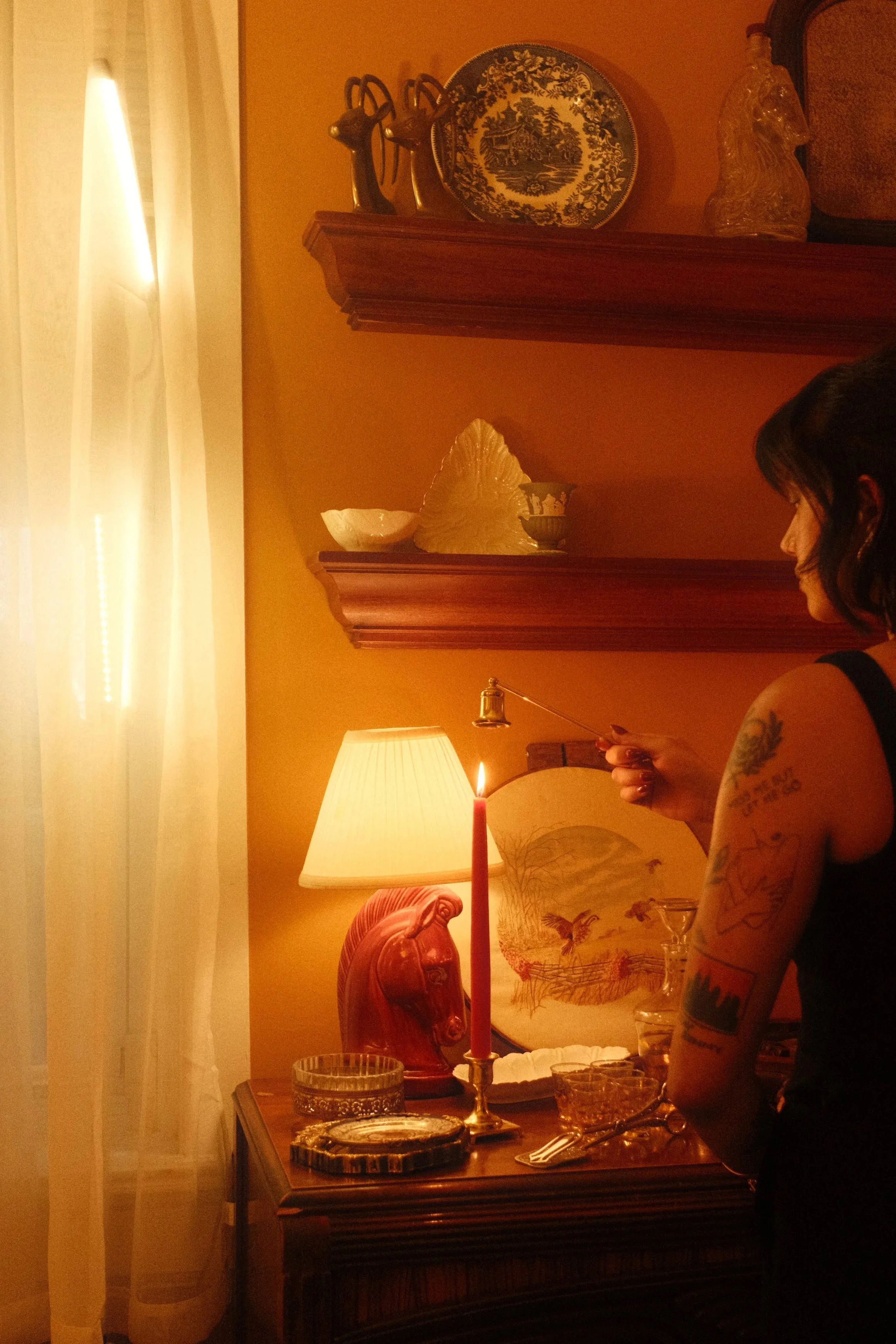 A woman with tattoos on her arm is standing by a table with a lit candle, glassware, and decorative items in a dimly lit room with warm lighting, a lamp, and artwork on the wall.