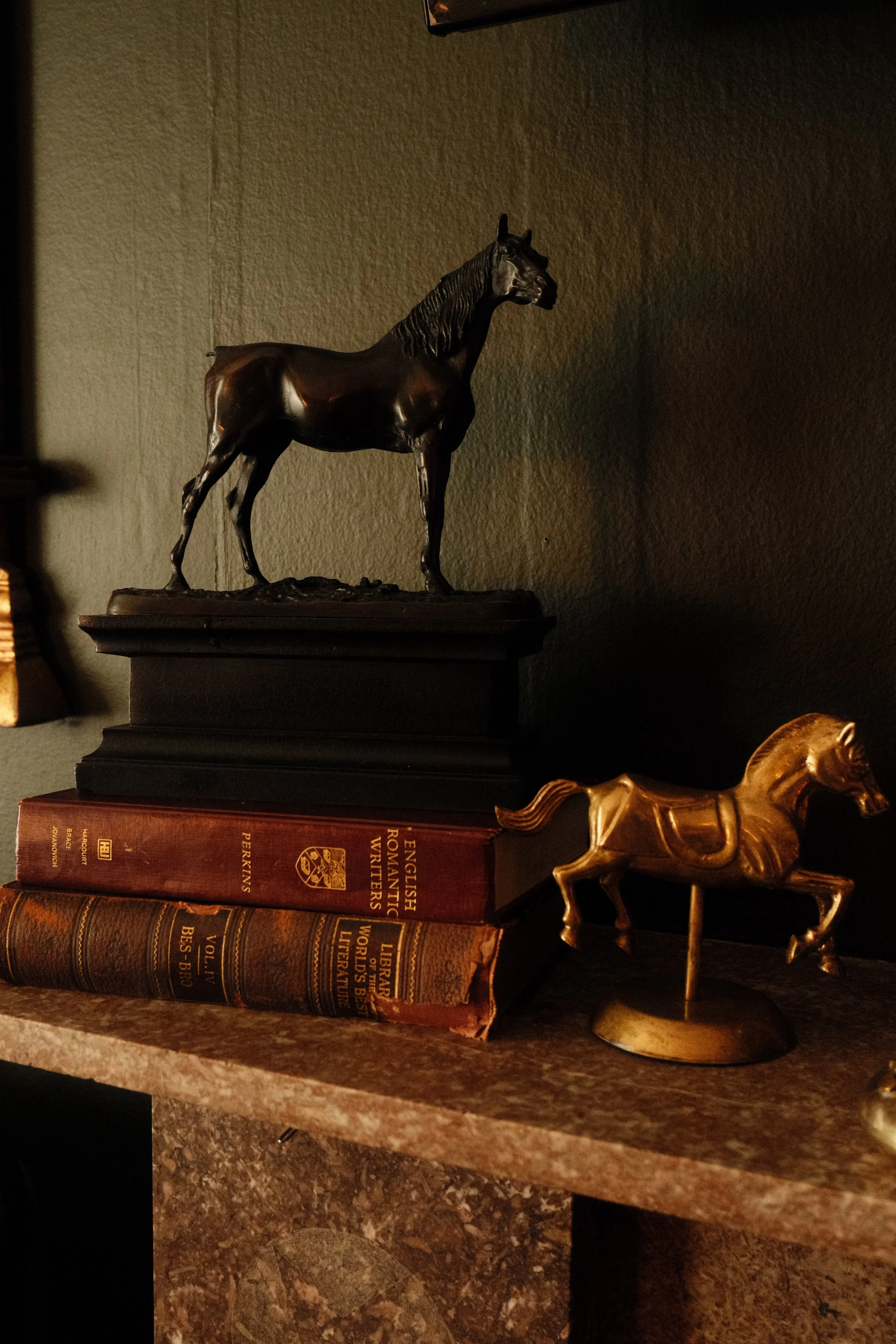 A bronze horse statue and a smaller gold horse statue displayed on a brown marble shelf alongside a set of three large old books with gold lettering.