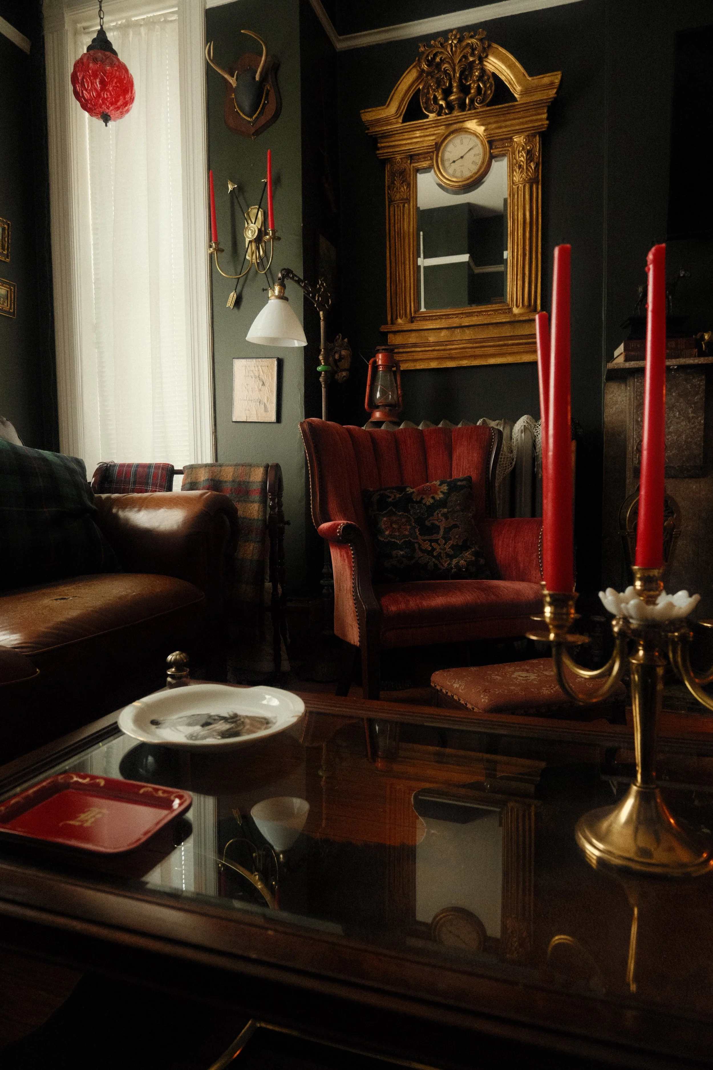 A cozy vintage living room with dark green walls, a gold ornate mirror with a clock, a wall-mounted deer head, red candles, and classic furniture including a leather armchair, a red upholstered chair, and a glass-topped coffee table with a decorative plate and a tray.